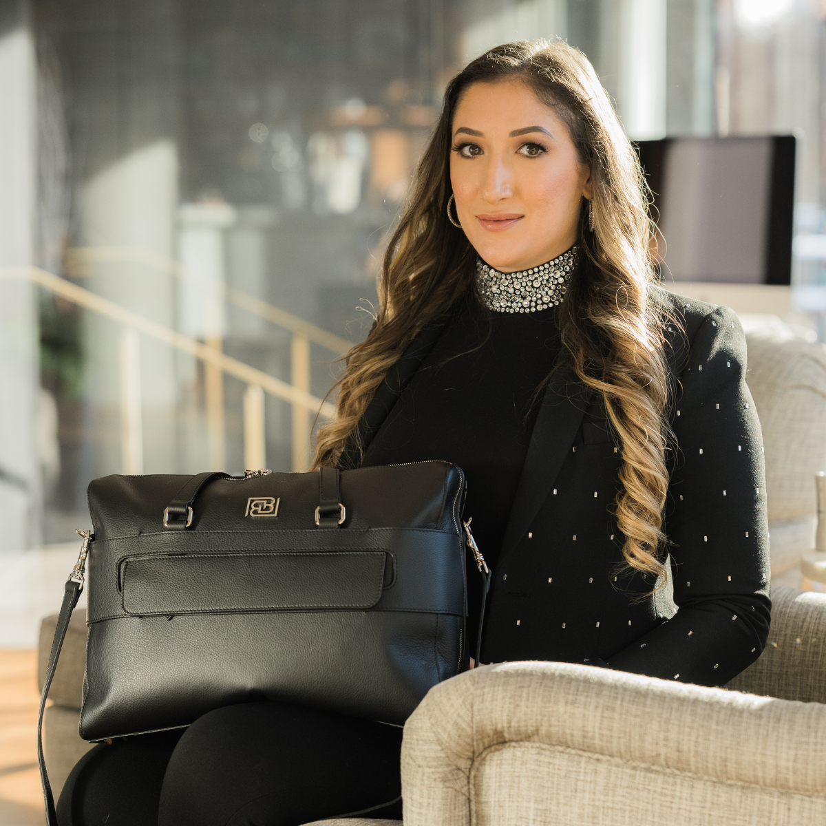 A woman with long wavy hair is sitting indoors, holding a black leather bag on her lap. She is wearing a black outfit with a sparkling, rhinestone-embellished collar and small decorative details on her blazer. She has hoop earrings and is looking at the camera with a slight smile.