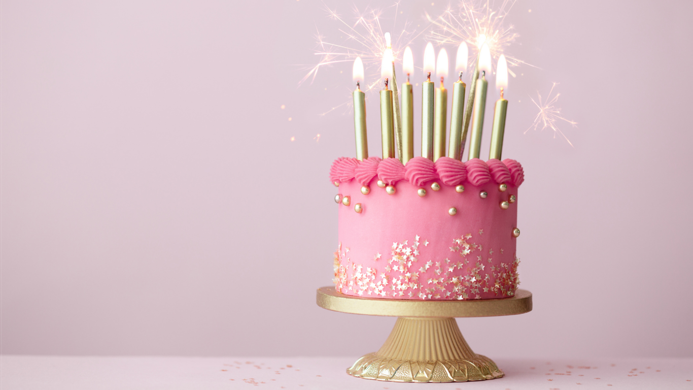 A pink birthday cake with gold and silver decorations, topped with lit candles and sparklers, sitting on a gold cake stand against a pink background.