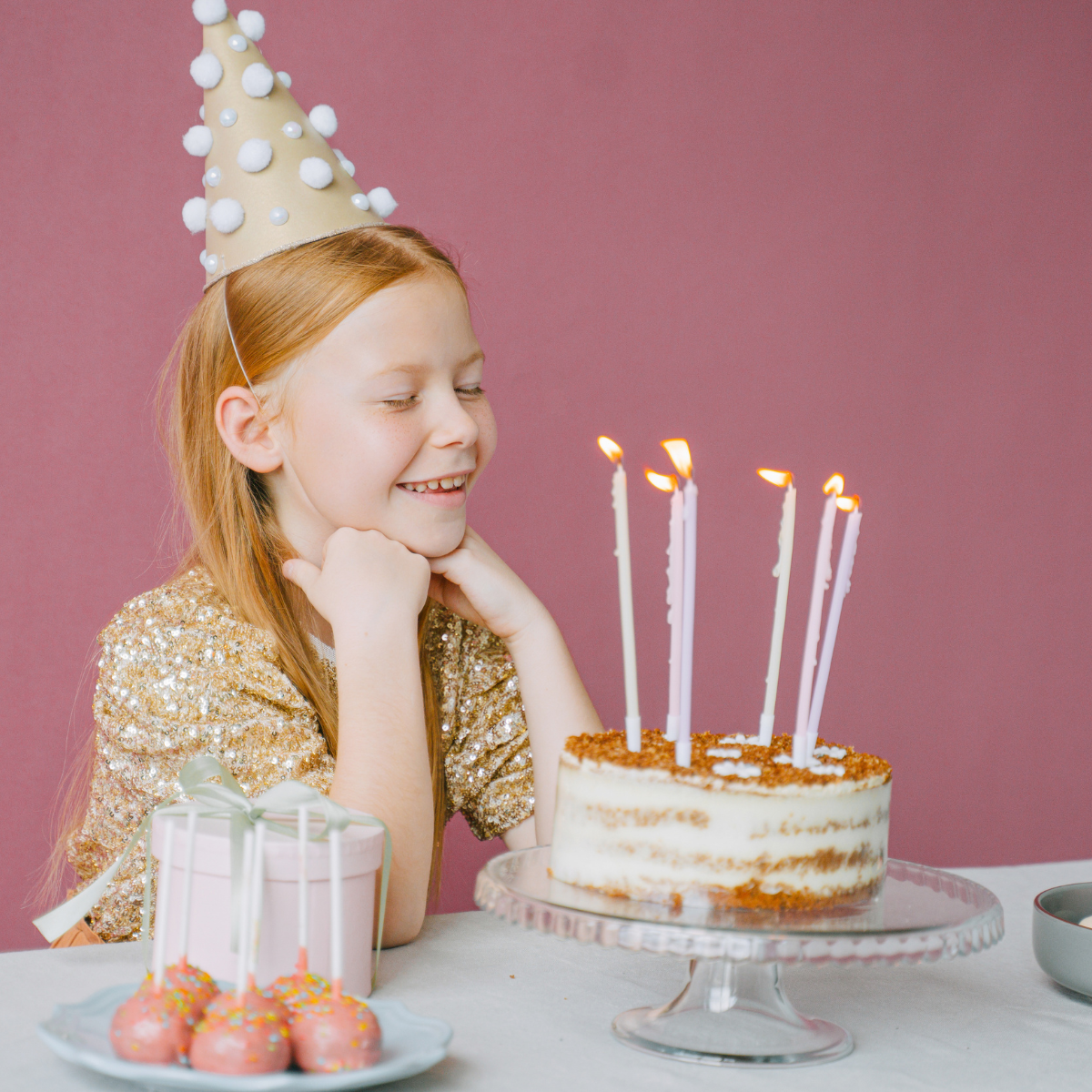 A young girl with red hair, wearing a party hat and a gold sequined top, sits at a birthday celebration with a cake with lit candles in front of her. She is smiling with her chin resting on her hands.