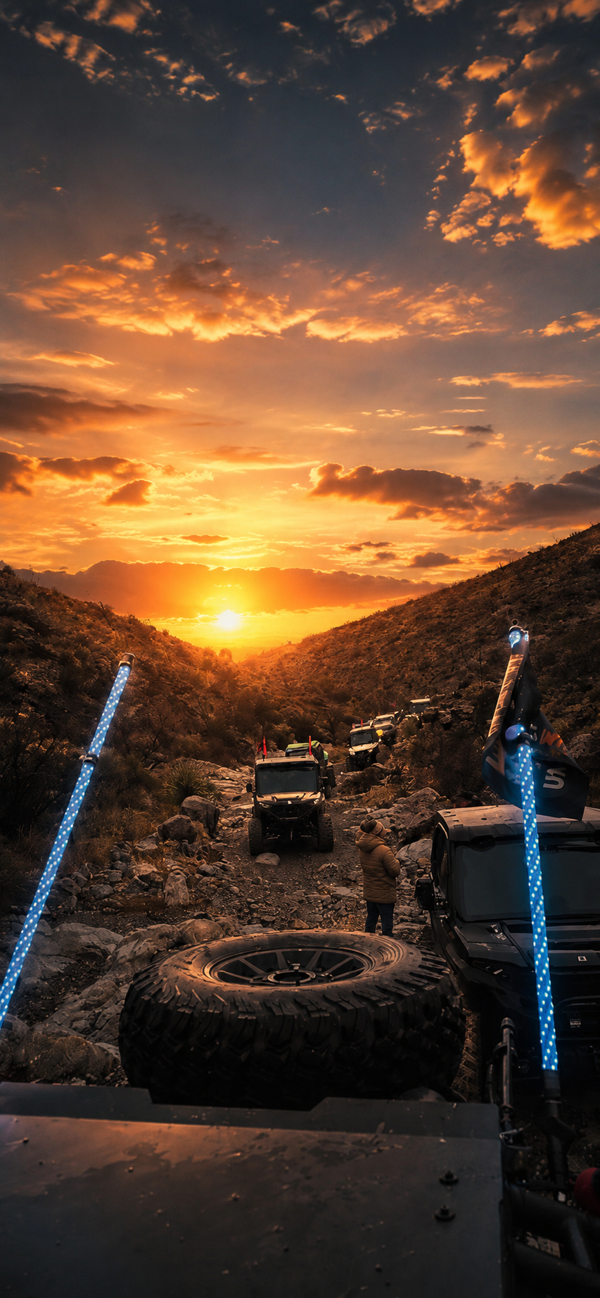 Off-road vehicles parked on a rocky trail in a canyon during sunset, with dramatic clouds in the sky and a person standing near a vehicle.