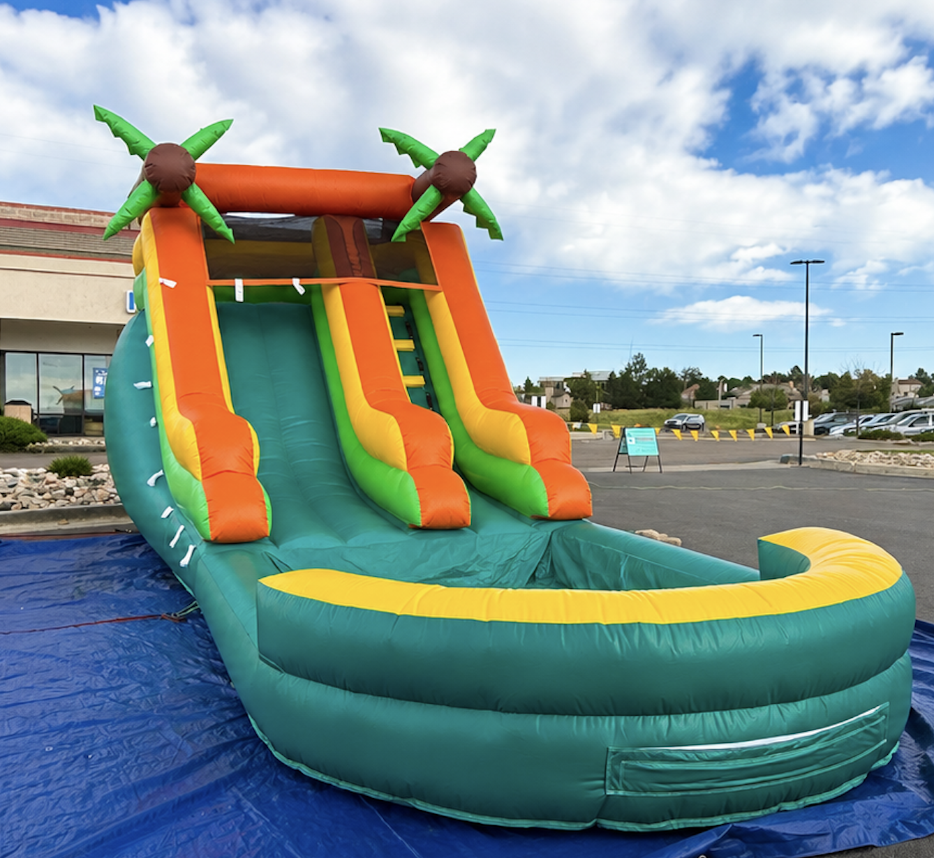 Colorful inflatable water slide with two orange slides, green and yellow accents, palm trees at the top, set up on a blue tarp in an outdoor parking lot with a store building and clear sky in the background.
