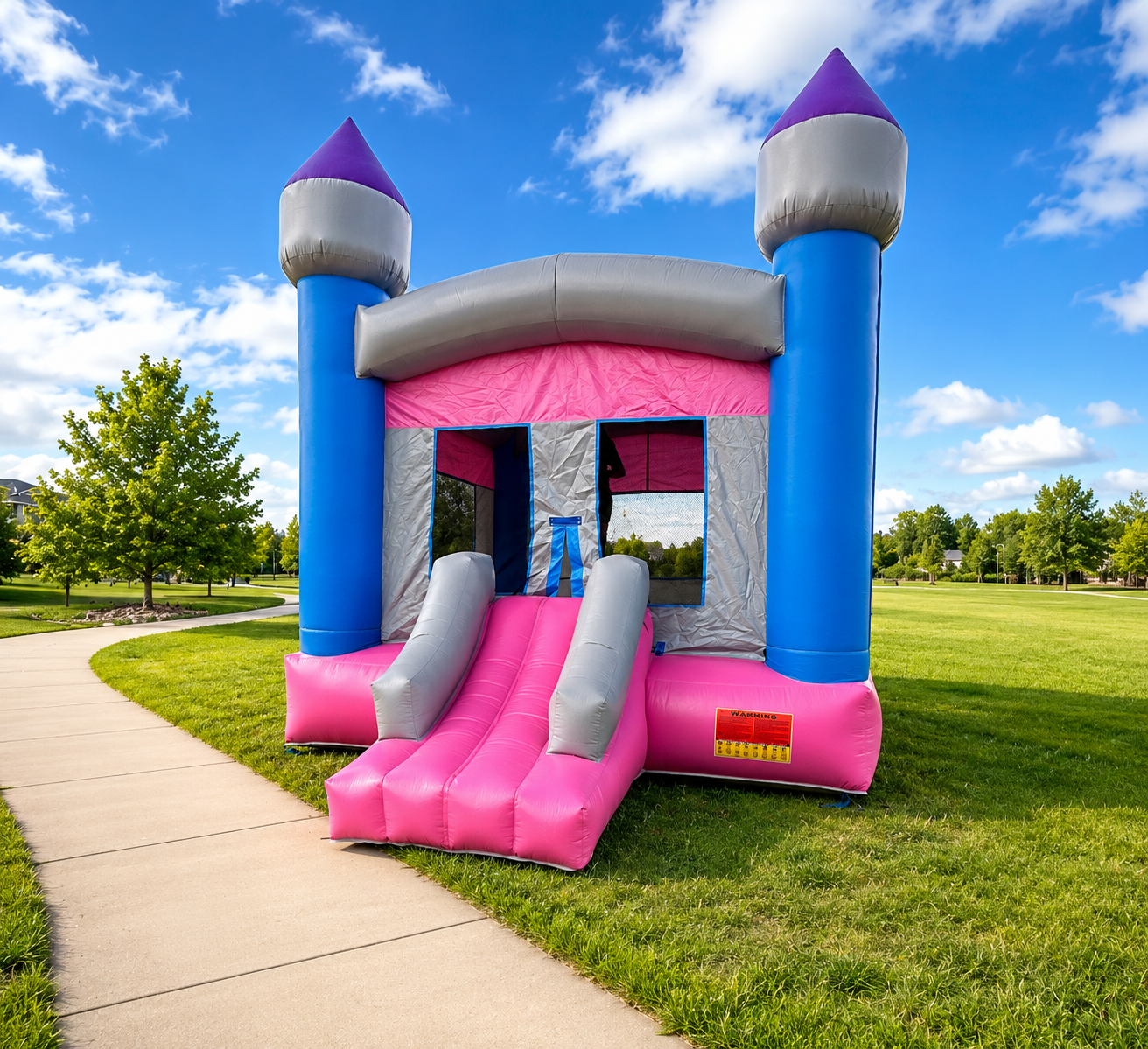 Colorful inflatable bounce house castle with pink and gray slides, blue towers with purple cones, set in a grassy park on a sunny day.