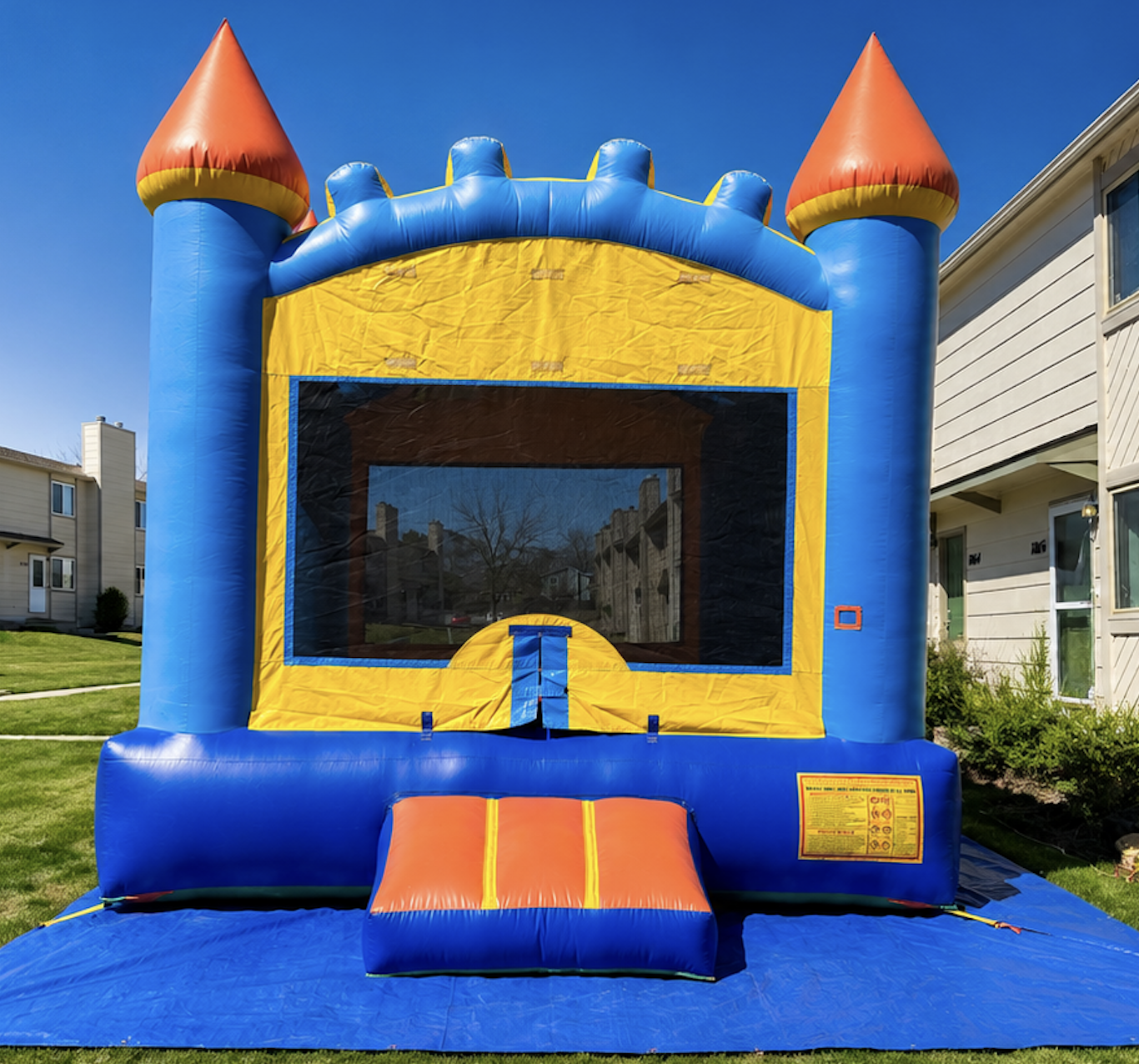 Colorful inflatable bounce house designed like a castle with blue, yellow, and orange sections, set up on a grassy area near residential buildings on a sunny day.