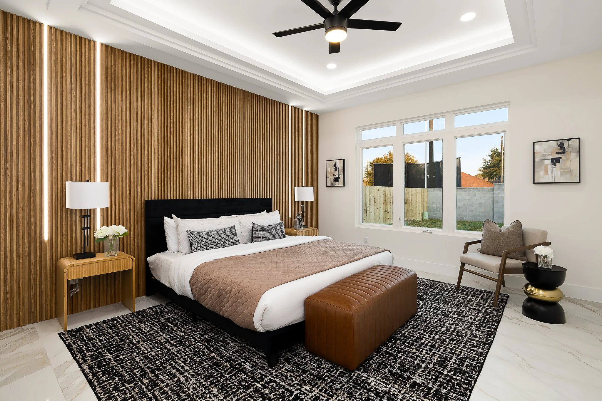 Modern bedroom with wooden accent wall, large windows, and contemporary furnishings including a bed with brown and white bedding, black headboard, side tables with lamps, gray armchair, and abstract wall art.