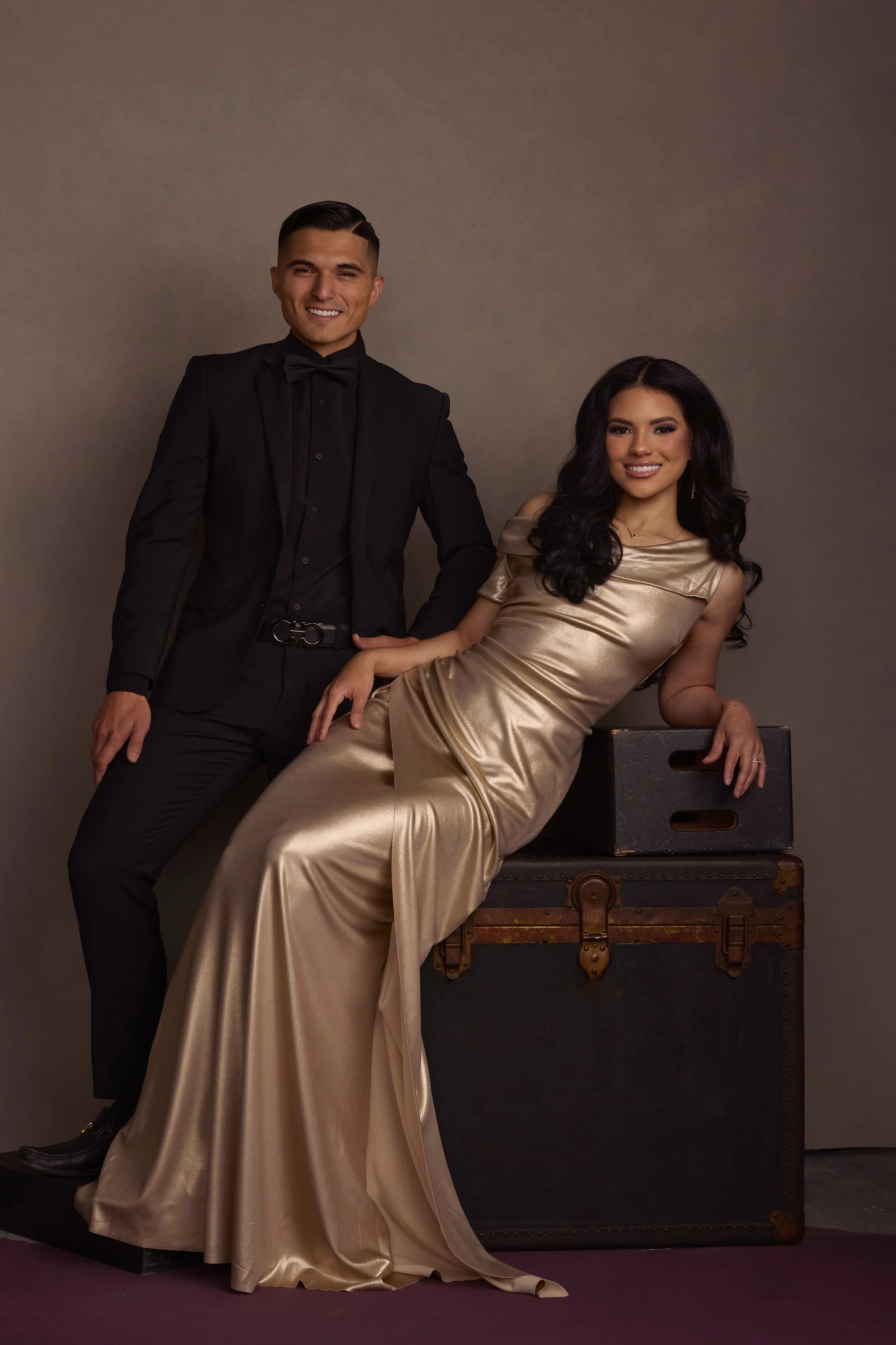 A young man in a black suit with a bow tie standing and a young woman in a gold satin gown sitting on a vintage trunk against a plain background.