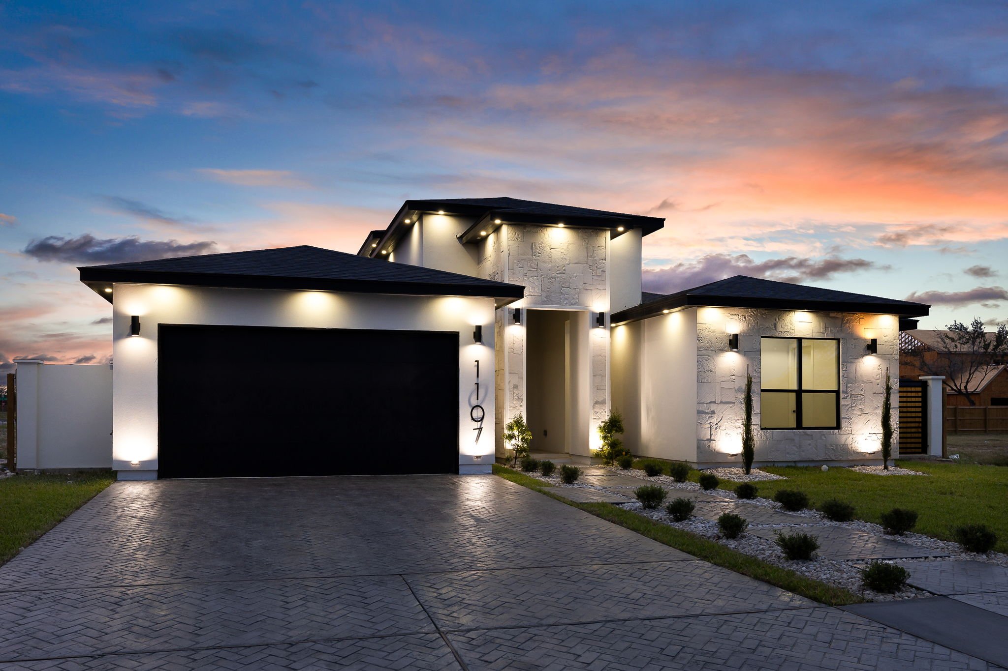 Modern house with white exterior walls, black roof, and illuminated outdoor lighting at sunset. The house features a garage with a black door, a front entryway, and landscaped yard with small plants and trees. The driveway is paved with brick-like patterns.