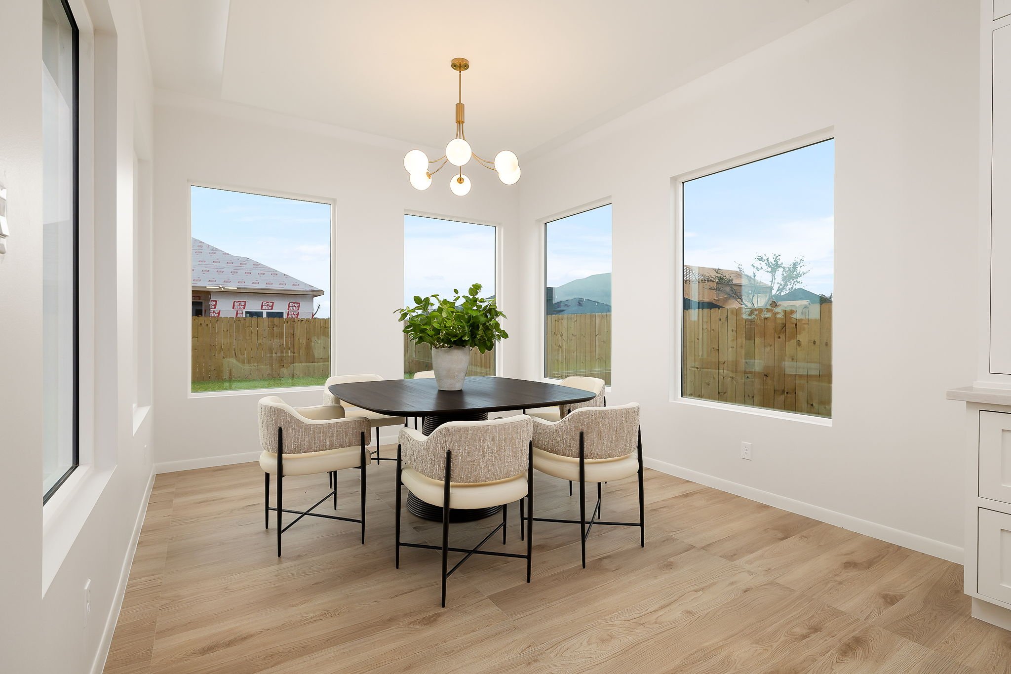 Modern dining room with six beige upholstered chairs around a dark round table, a white pot with a green plant, large windows, white walls, wooden flooring, and a contemporary chandelier.