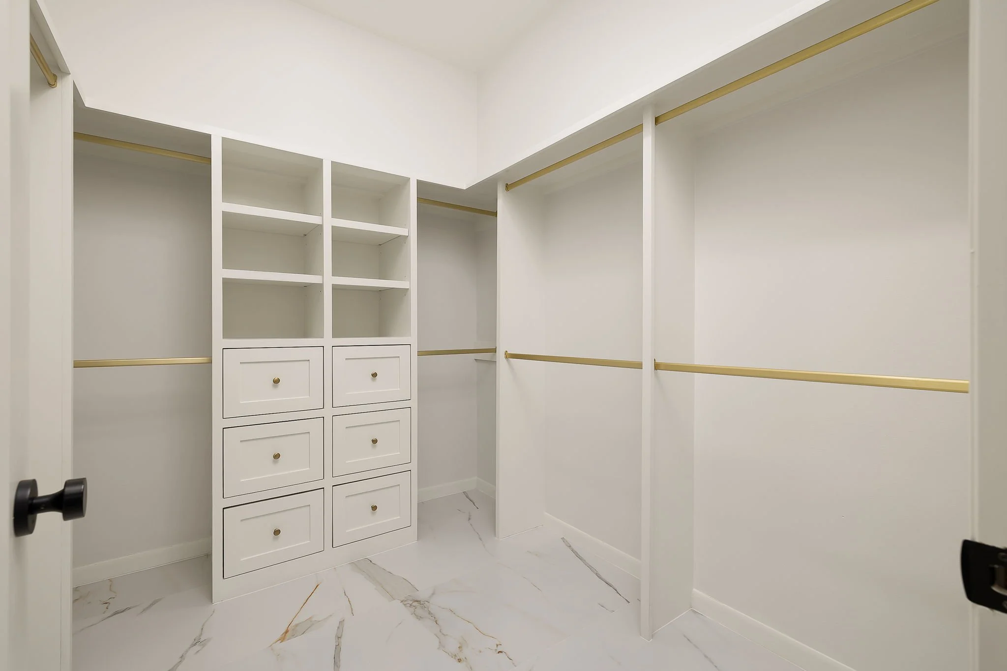 Empty walk-in closet with white shelves and drawers, gold hanging rods, marble floor, and a door handle visible on the left.