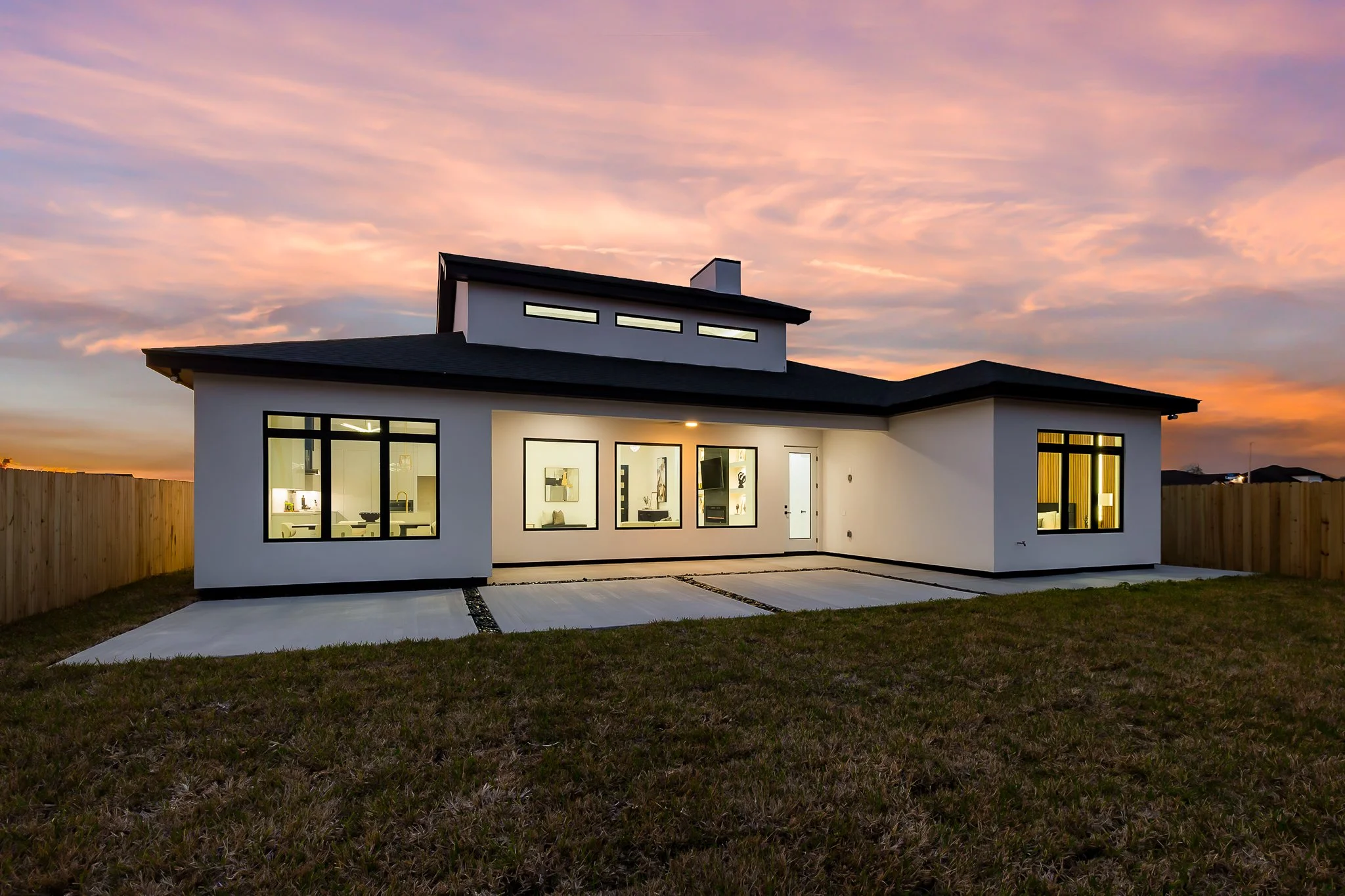 Modern white house with large windows illuminated from inside, set against a colorful evening sky with pink and purple clouds, surrounded by a wooden fence and a grassy yard.