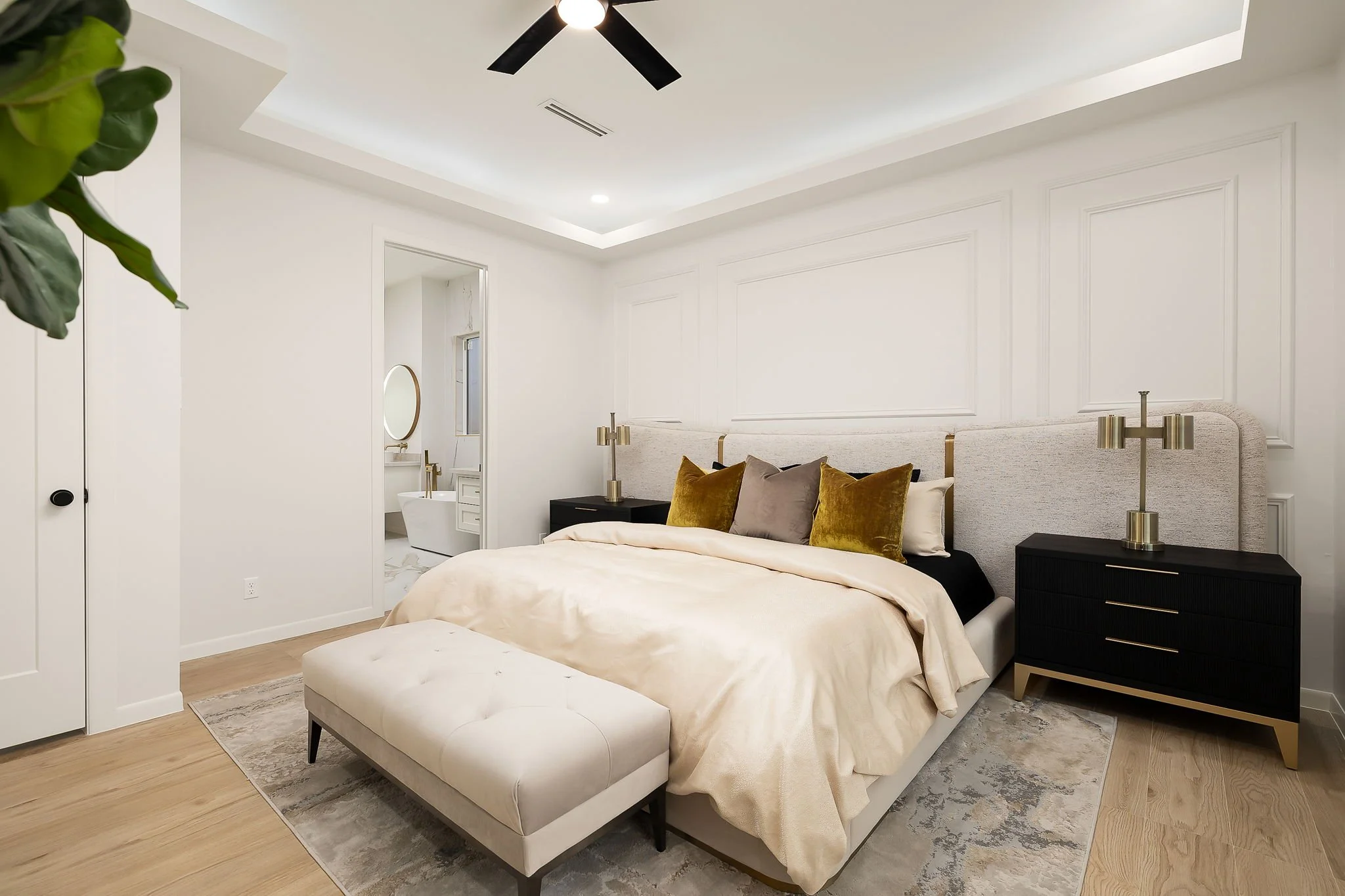 A modern bedroom with a large bed featuring a beige bedspread and multiple pillows, black nightstands with gold accents, a plush bench at the foot of the bed, and a ceiling fan with black blades. There's a small hanging lamp on each side of the bed, a mirror visible in the background, and a plant partially visible on the left side.