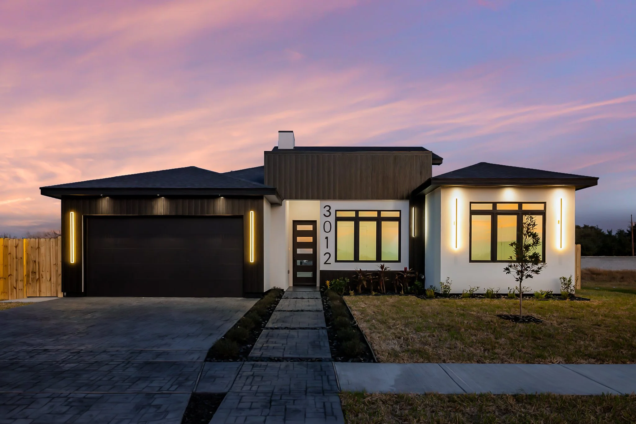 Modern single-story house with a dark roof, large front windows, a garage, and a small front yard with a young tree, illuminated exterior lighting, and a colorful evening sky in the background.