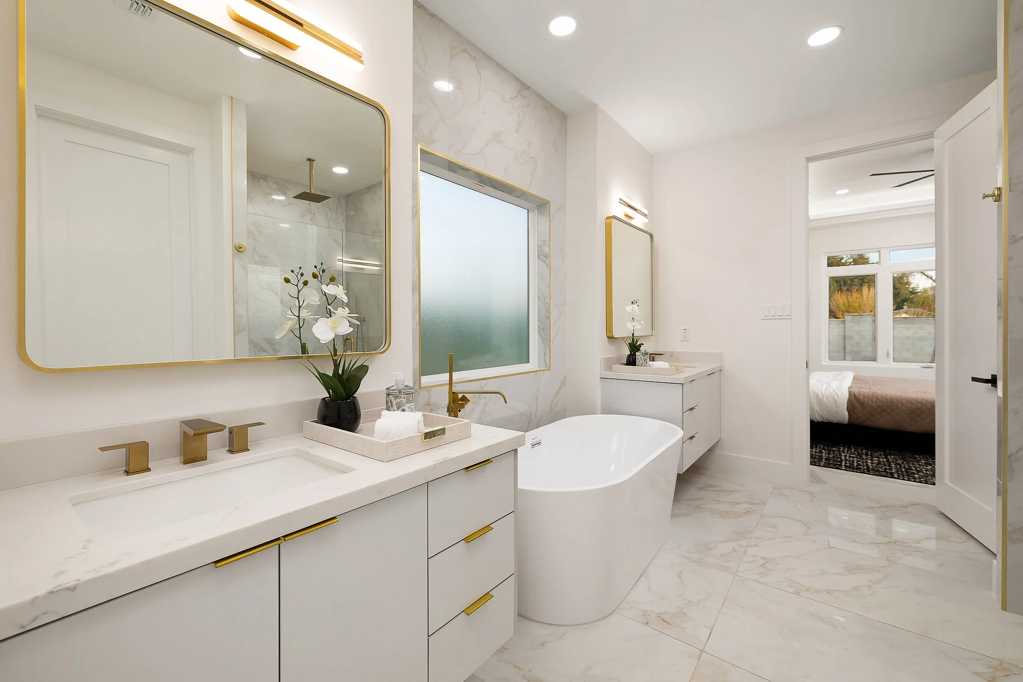 A modern bathroom with white marble countertops, gold accents, and a white freestanding bathtub. There are two large rectangular mirrors framed in gold, a black vase with white orchids, and a vanity area with gold faucet fixtures. The room has a large frosted window, recessed lighting, and a view of a bedroom with large windows and a bed.