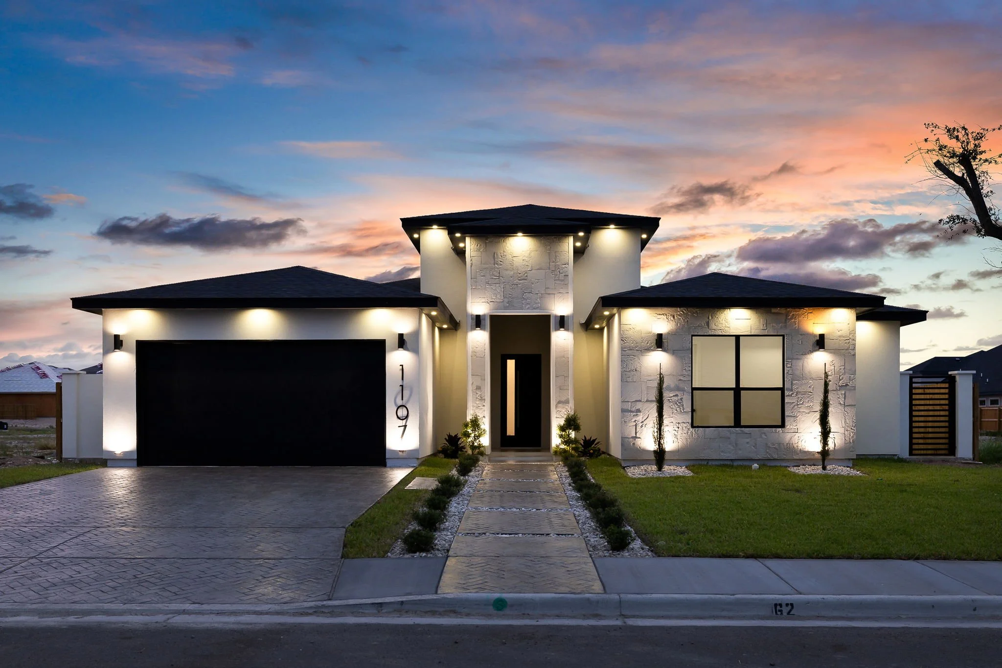 Modern two-story house with a white exterior, black accents, and outdoor lighting at dusk. Features a paved driveway, a small landscaped front yard with plants, and a pathway leading to the entrance. The sky is partly cloudy with a colorful sunset.