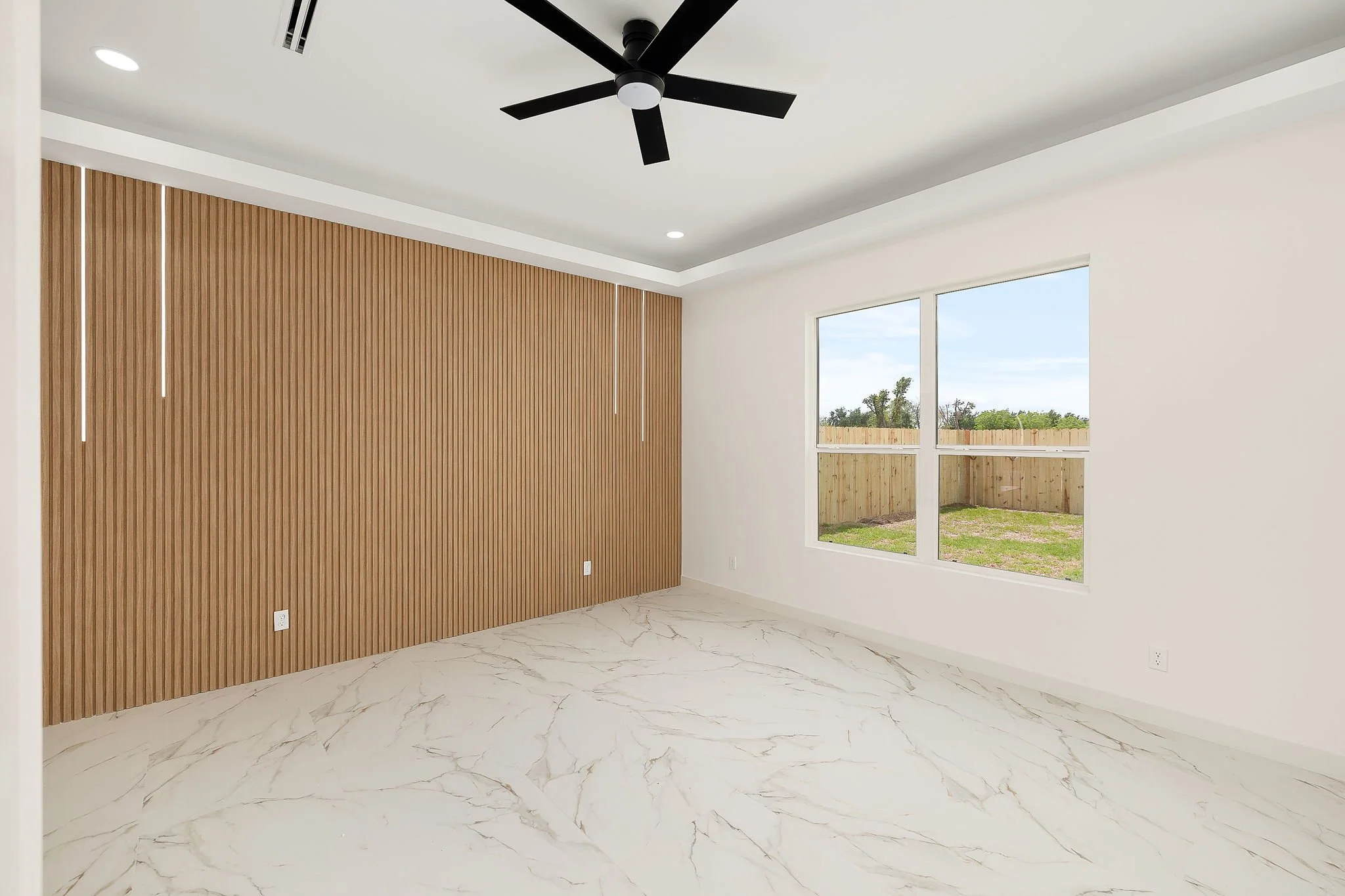 Empty living room with white walls, a wooden slat accent wall, large windows showing an outdoor yard, marble-patterned flooring, recessed and ceiling fan lighting.