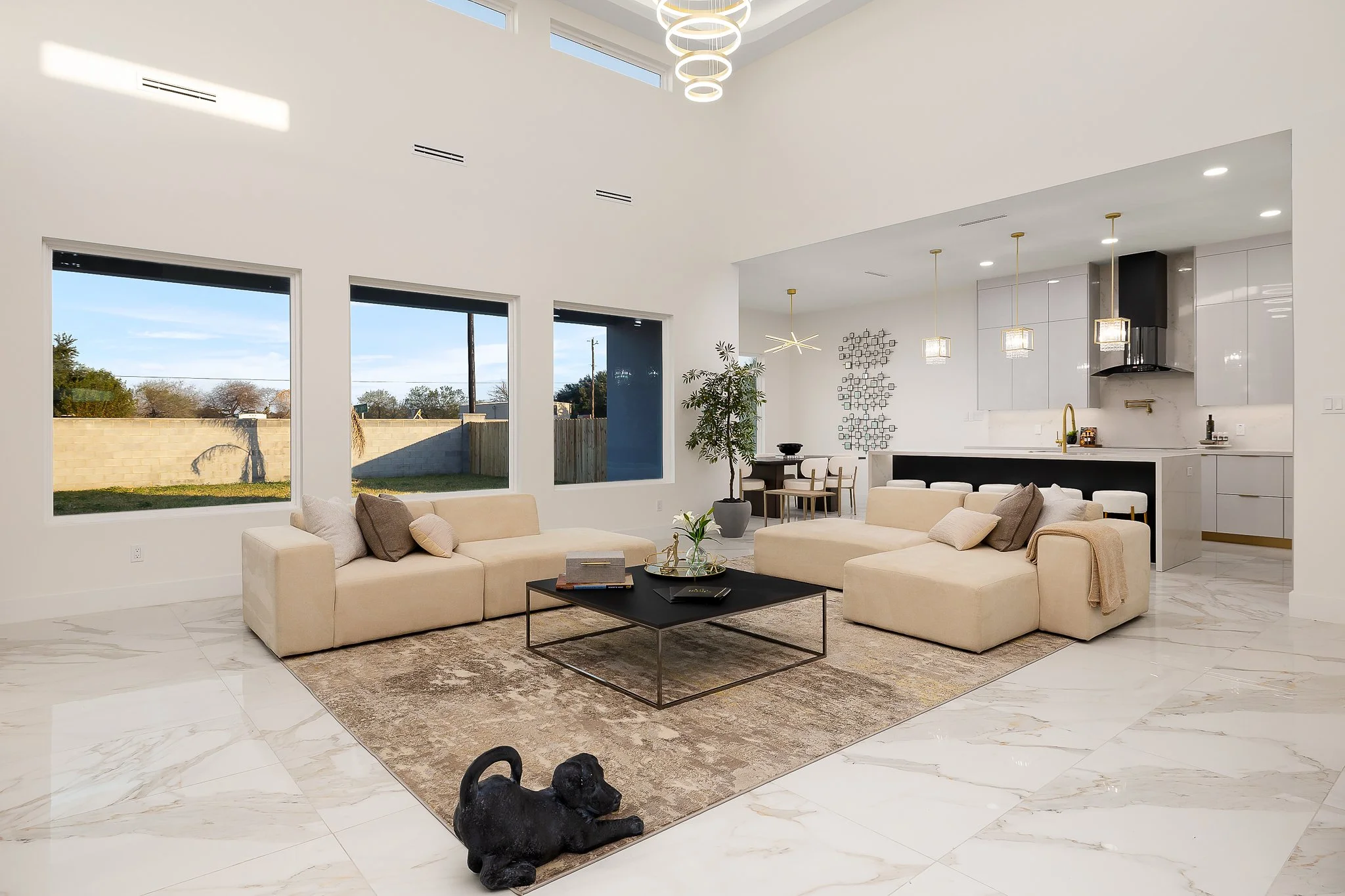 Modern living room with beige sofas, a black coffee table, a decorative rug, and large windows showing an outdoor yard. White walls, marble flooring, and minimalist lighting fixtures, with a black dog sculpture on the floor.
