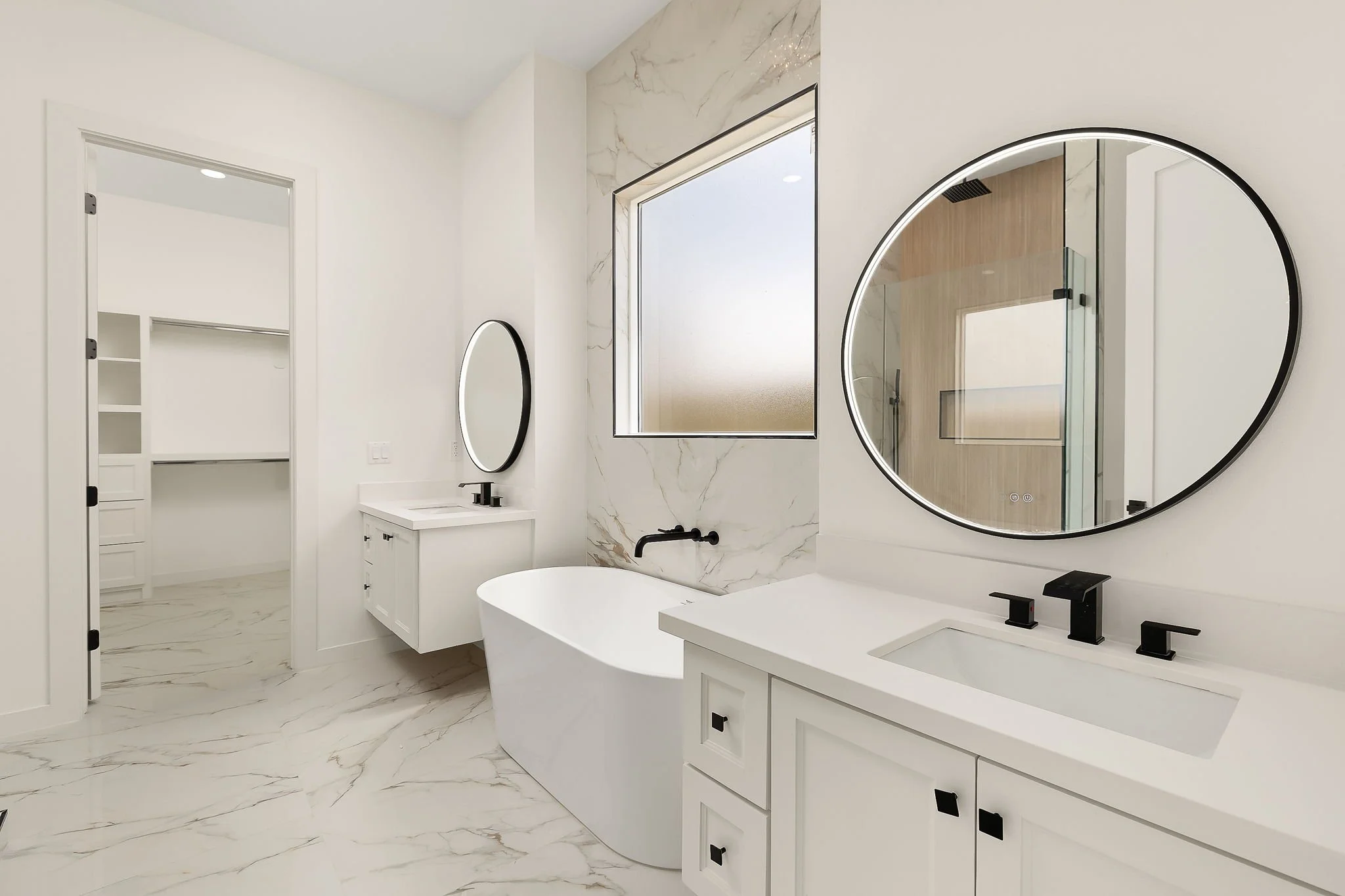 Modern bathroom with white cabinetry, marble floors, and a large round illuminated mirror above the sink.
