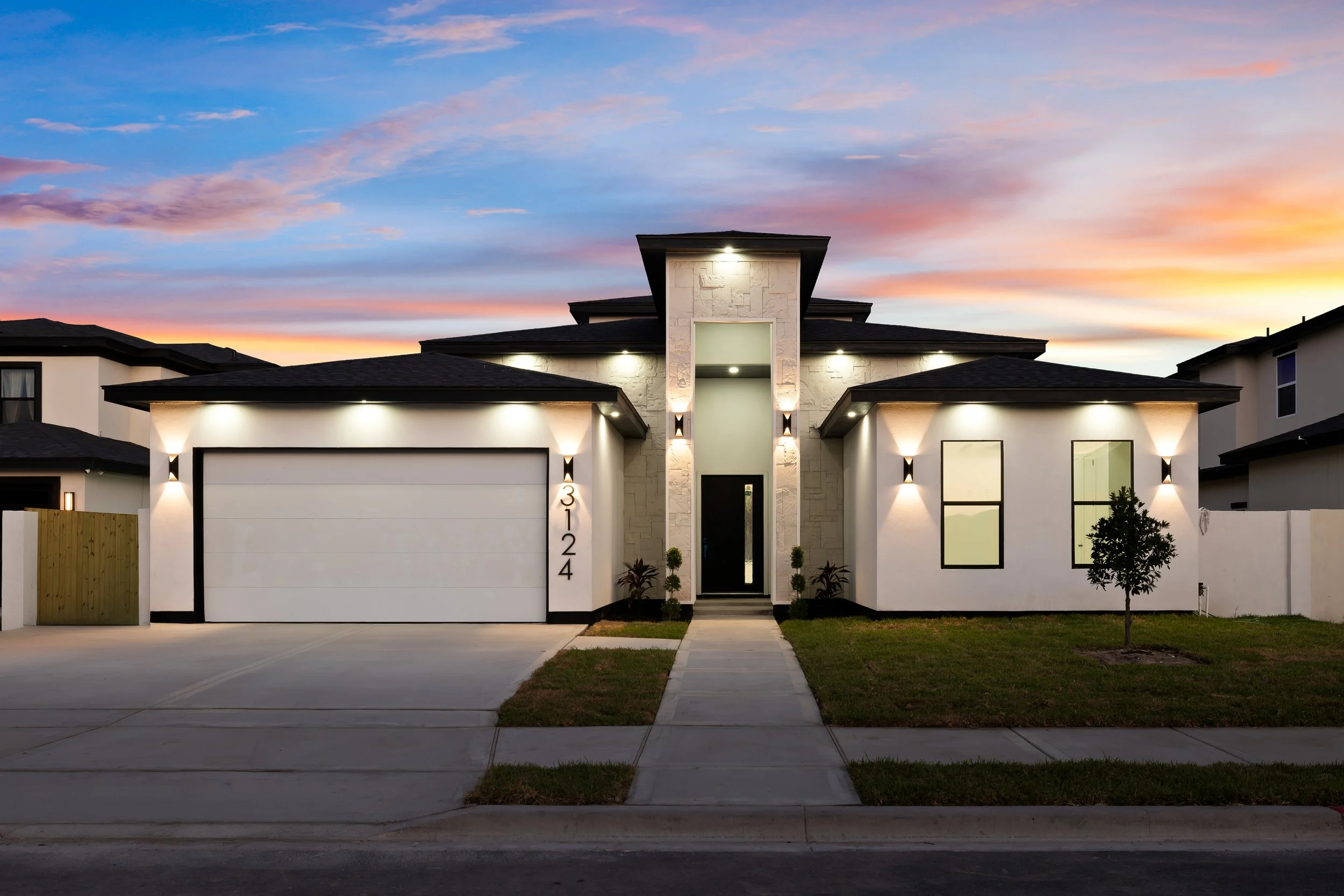 Modern single-family house with a white facade, black roof, and a two-car garage, illuminated by exterior lights, with a small front yard and a tree, during sunset.
