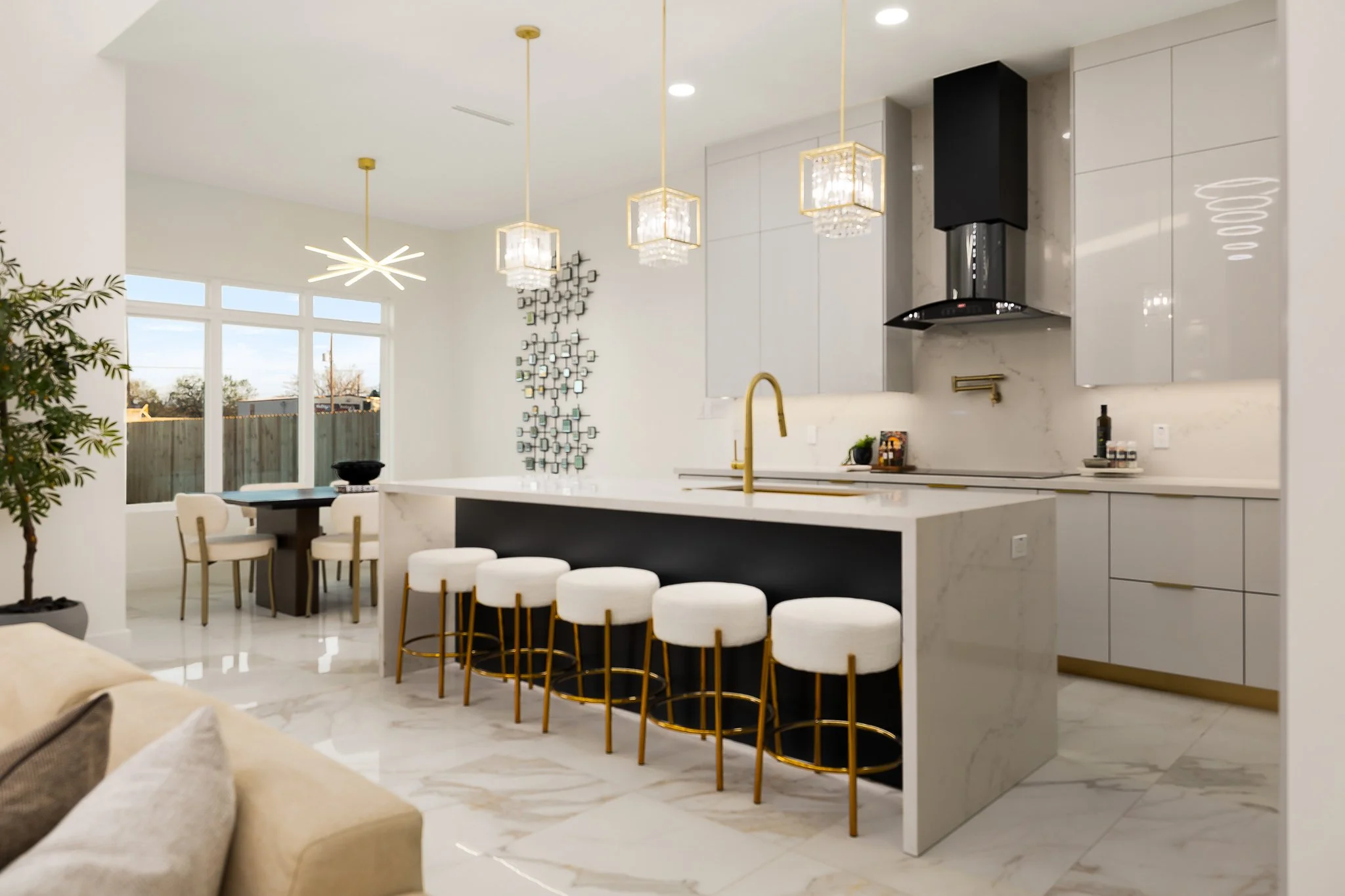Modern kitchen and dining area with white cabinets, marble island with gold accents, chandelier, and large windows showing outdoor scenery.