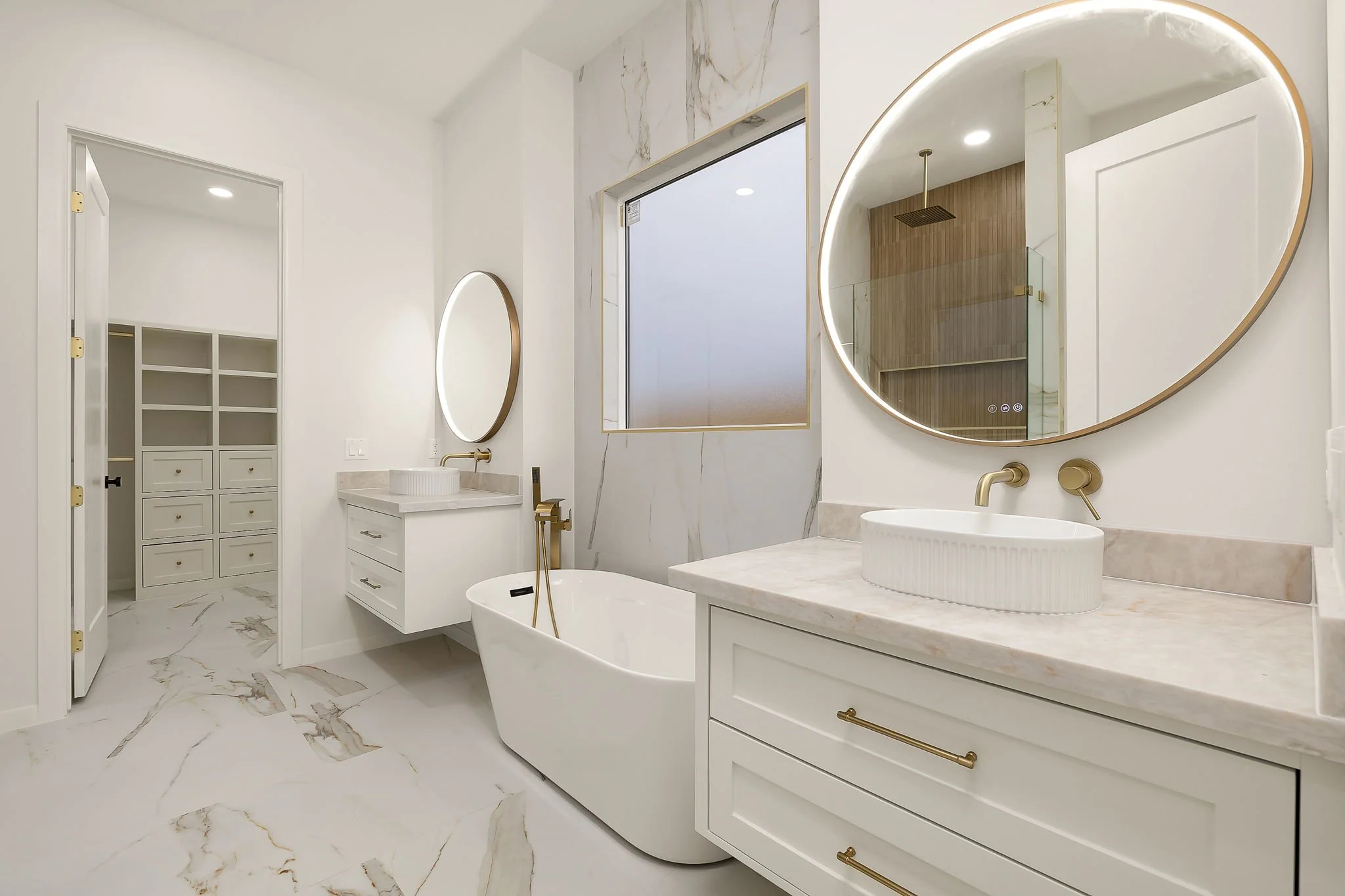 Modern bathroom with white vanity, oval mirror with gold trim, round vessel sink, gold fixtures, marble floor and walls, and a walk-in shower with glass door and wood panel ceiling.