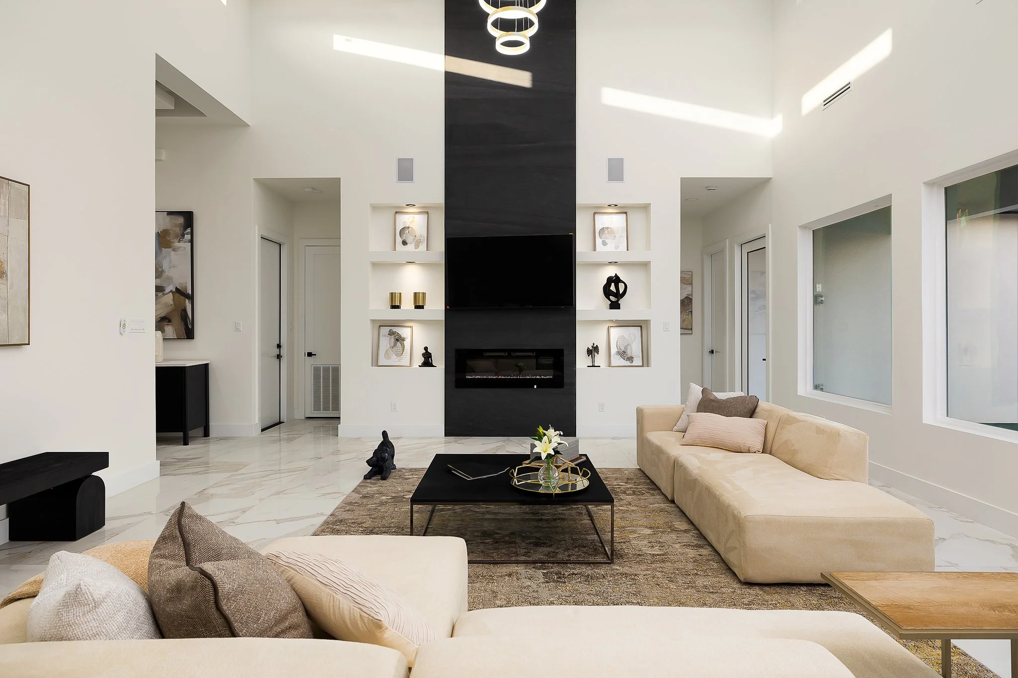 Modern living room with white walls, a black fireplace, and built-in shelves with artwork and decor. Light-colored sofas, a dark coffee table with flowers, and abstract artwork on the walls. Large windows and a high ceiling with circular light fixtures.