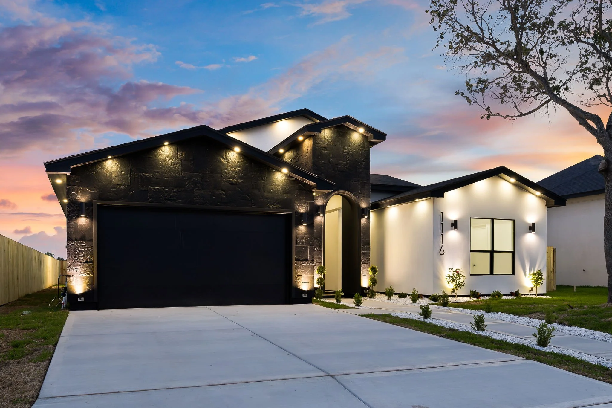 Modern house with dark stone and white exterior, illuminated by outdoor lights at dusk, with a large tree to the side and a sunset sky.