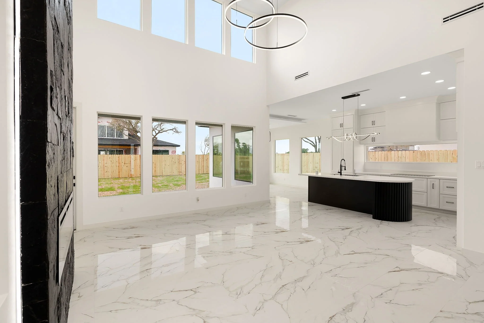 Modern kitchen and living area with white walls, large windows, and marble flooring. Features a black kitchen island and pendant lighting.