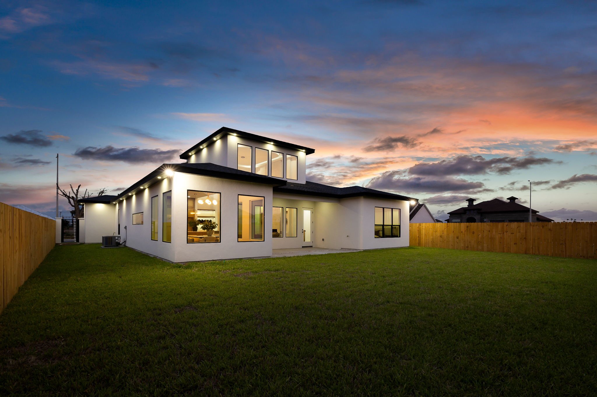Modern two-story house with large windows, surrounded by a grassy backyard with a wooden fence, during sunset with a colorful sky and some clouds.