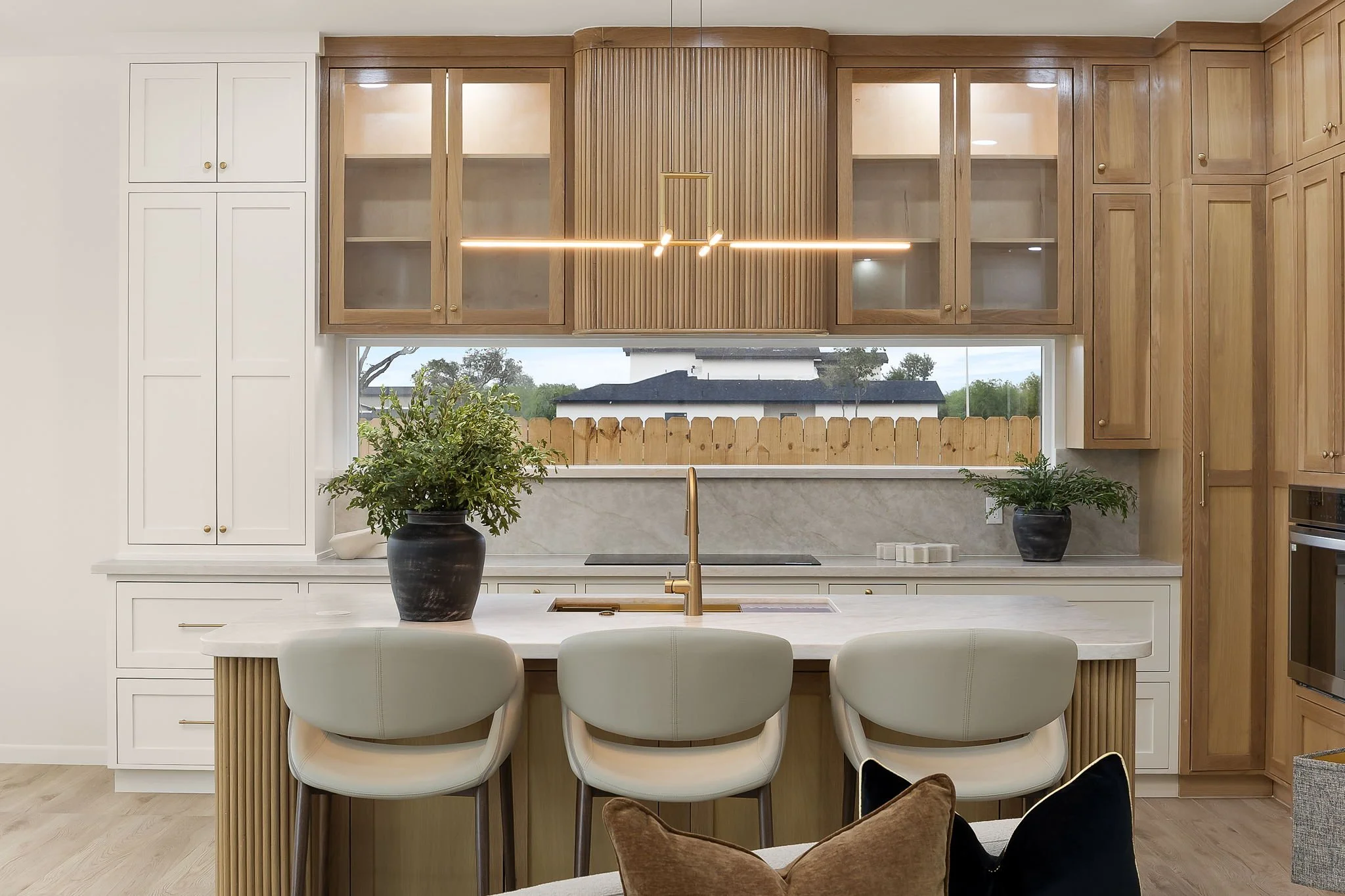 Modern kitchen with white and wood cabinetry, a large window overlooking a suburban yard, potted plants on the counter, and a sleek hanging light fixture.
