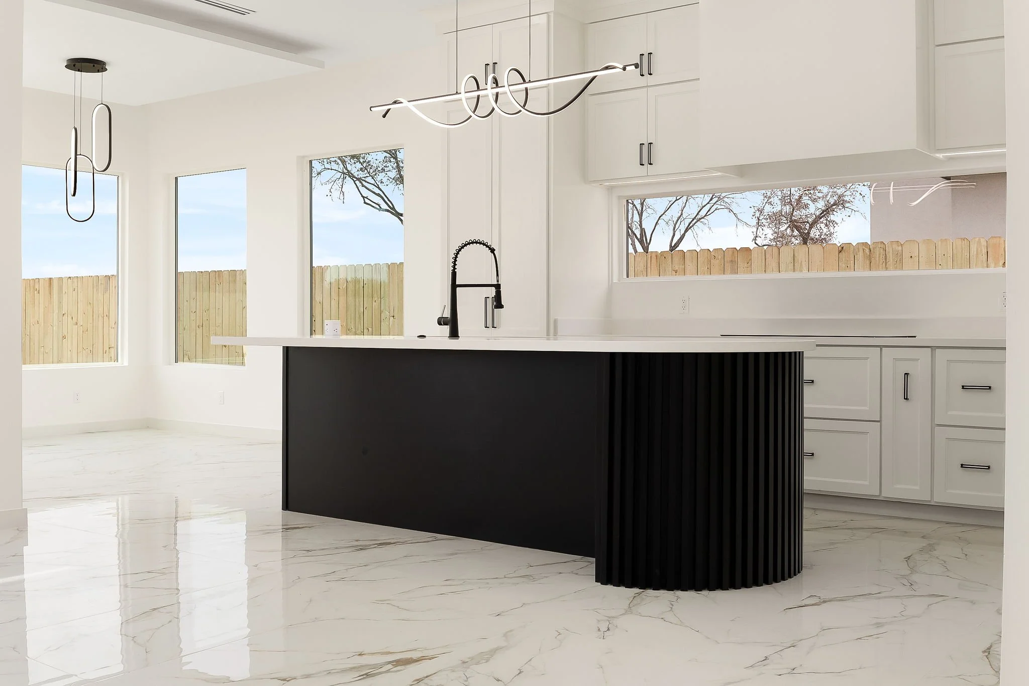 Modern kitchen with white cabinets, large windows showing trees outside, a black island with vertical paneling, a black faucet, and contemporary lighting fixtures.