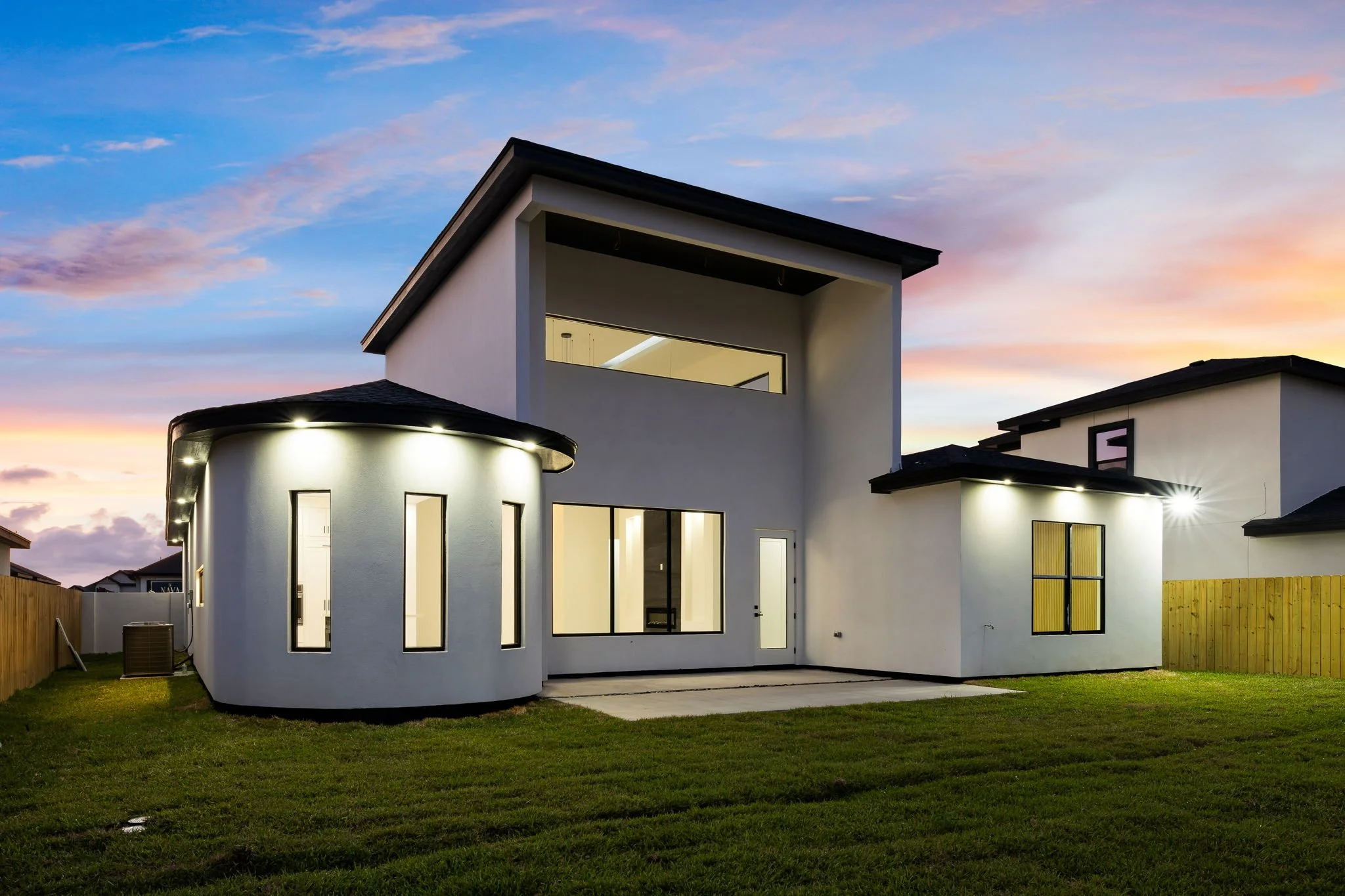 Modern two-story house with white exterior walls, large windows, and a flat roof, illuminated at dusk with a colorful sky in the background.
