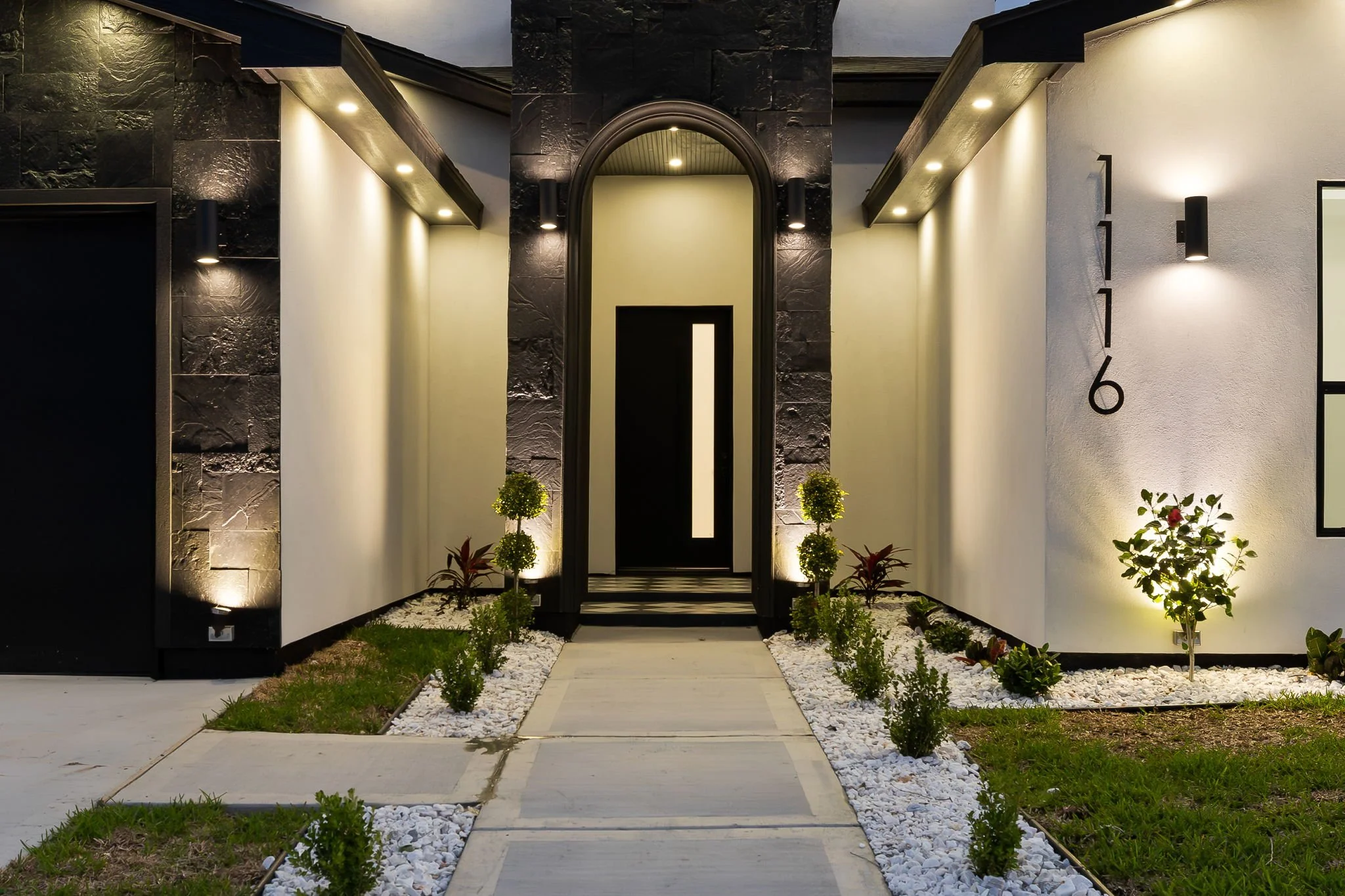Modern house entrance with stone and white wall, black archway, potted plants, and pathway lights