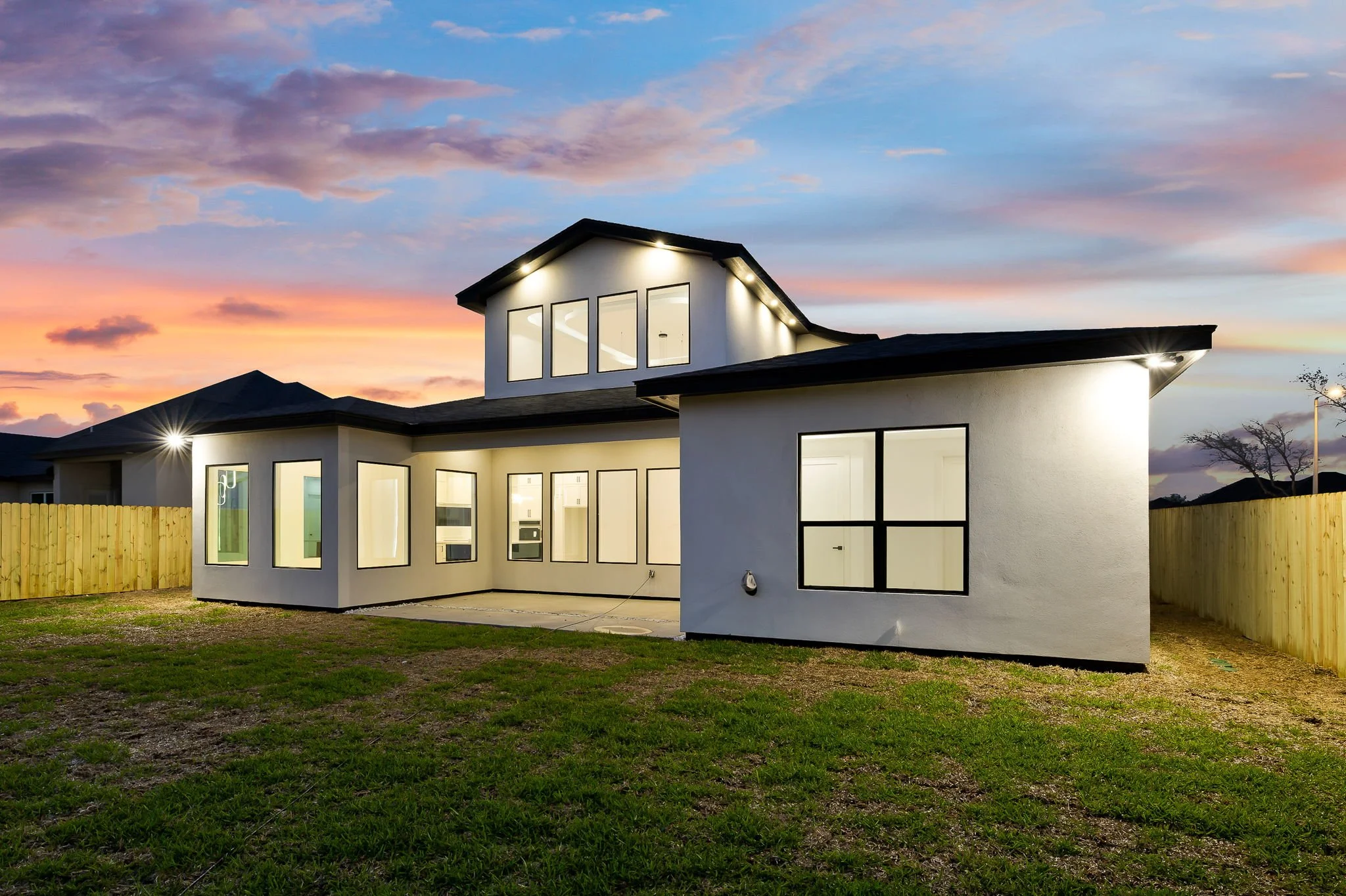 Modern white house with black trim and large windows, illuminated from inside, under a colorful evening sky, surrounded by a wooden fence and patchy grass yard.