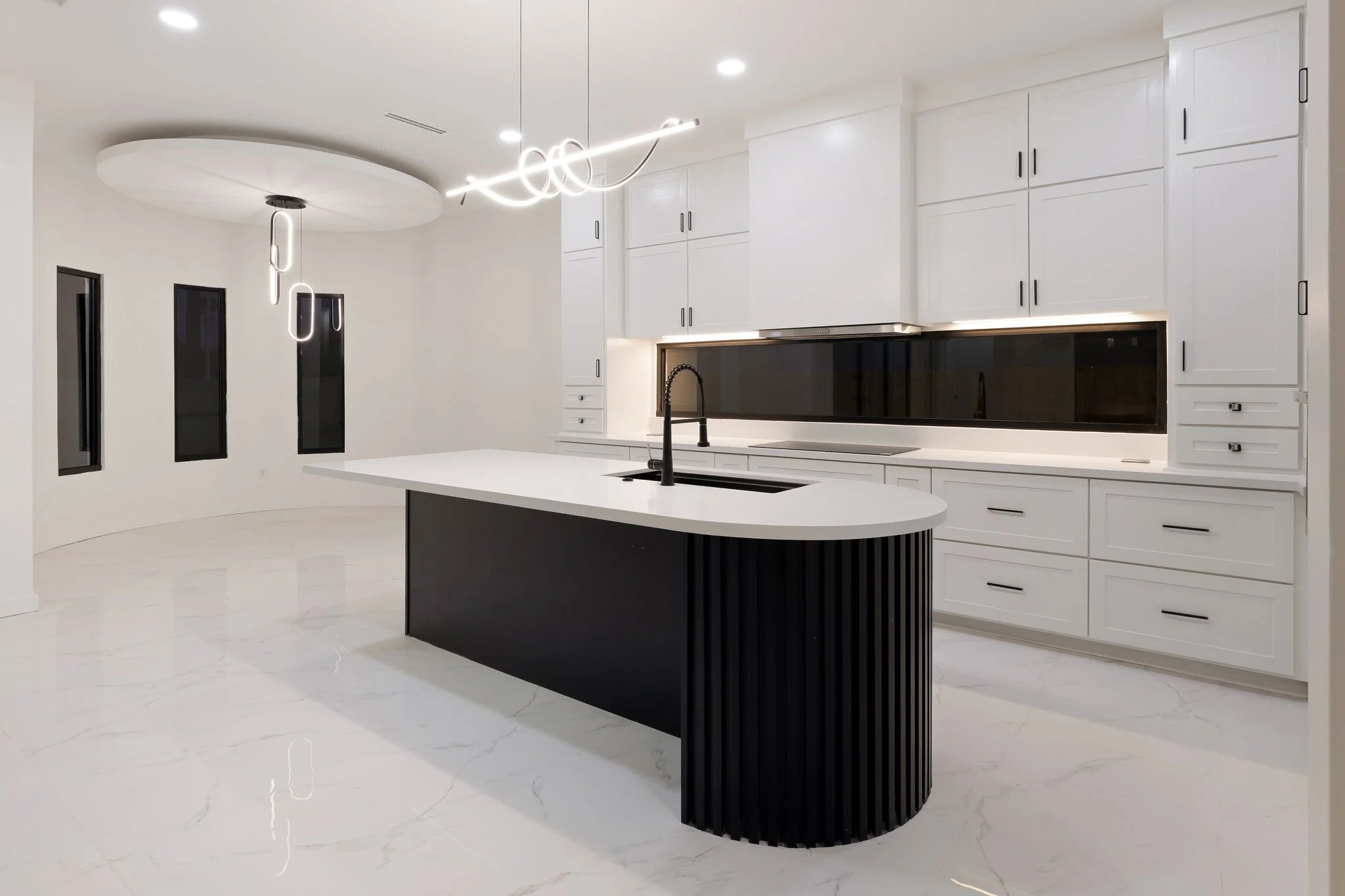 Modern kitchen with white cabinets, black countertop, black sink, island with black base, and stylish black pendant and linear lights, marble floor, and three narrow windows.