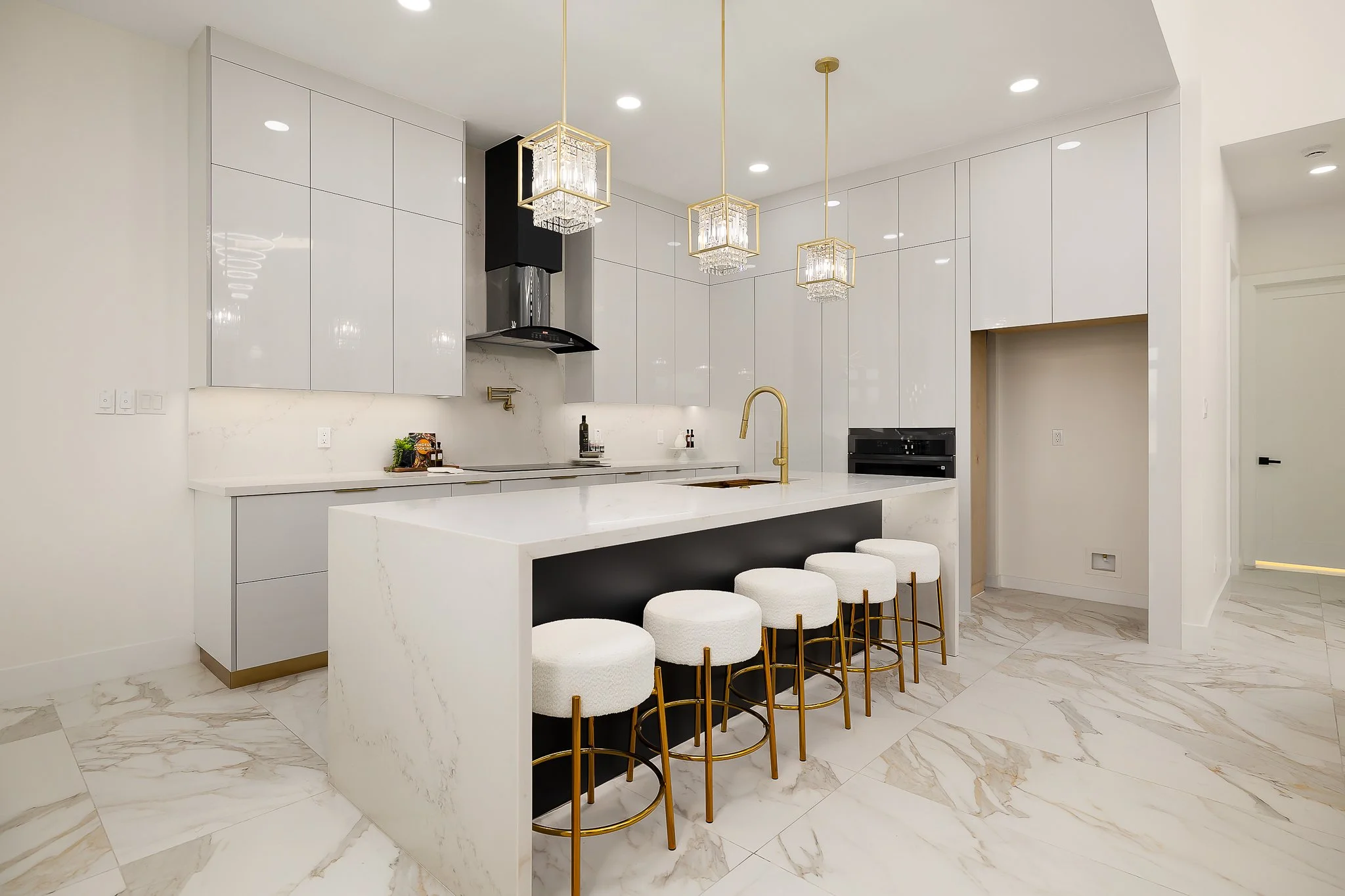 Modern white kitchen with marble flooring, a large island with bar stools, and gold accents. Hanging chandelier lights are suspended above the island, and built-in appliances are visible along the wall.
