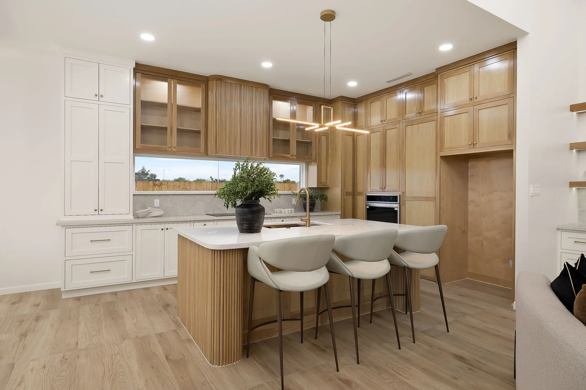 Modern kitchen with white and wooden cabinets, marble island with four white chairs, potted plant, large window, and minimalist lighting.