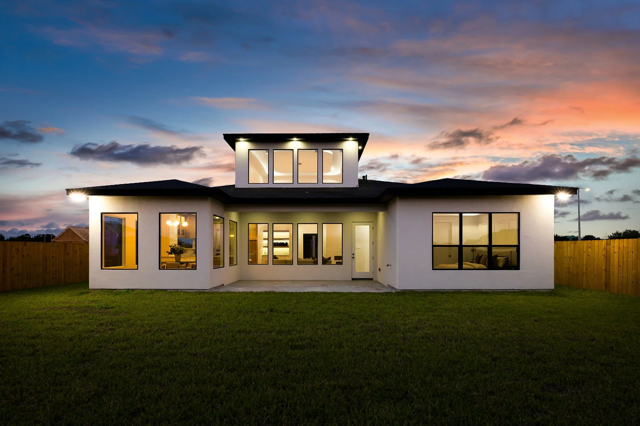 Modern two-story house with large windows, illuminated interior, and a dark roof, set against a colorful sunset sky.