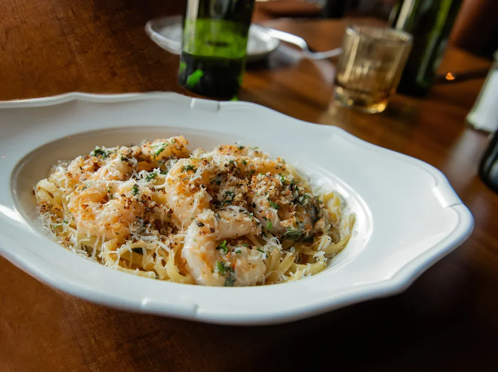 Fettuccine pasta with shrimp, garnished with herbs and grated cheese, served in a white oval dish on a wooden table.