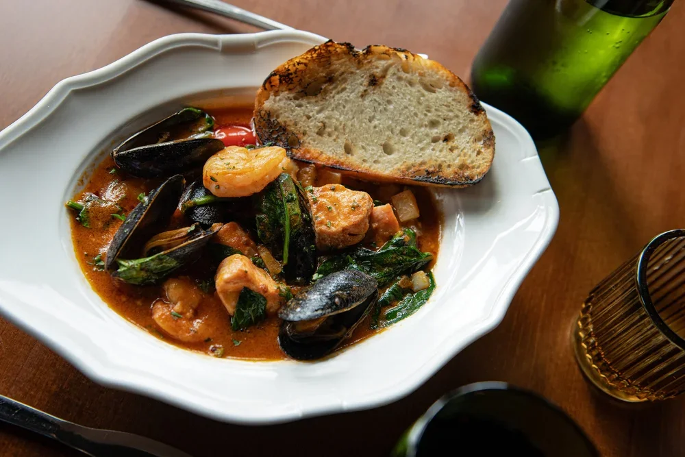 Seafood stew with mussels, shrimp, and vegetables served in a white bowl, with a slice of toasted bread on the side. A glass bottle of green drink and a coffee mug are on the table.