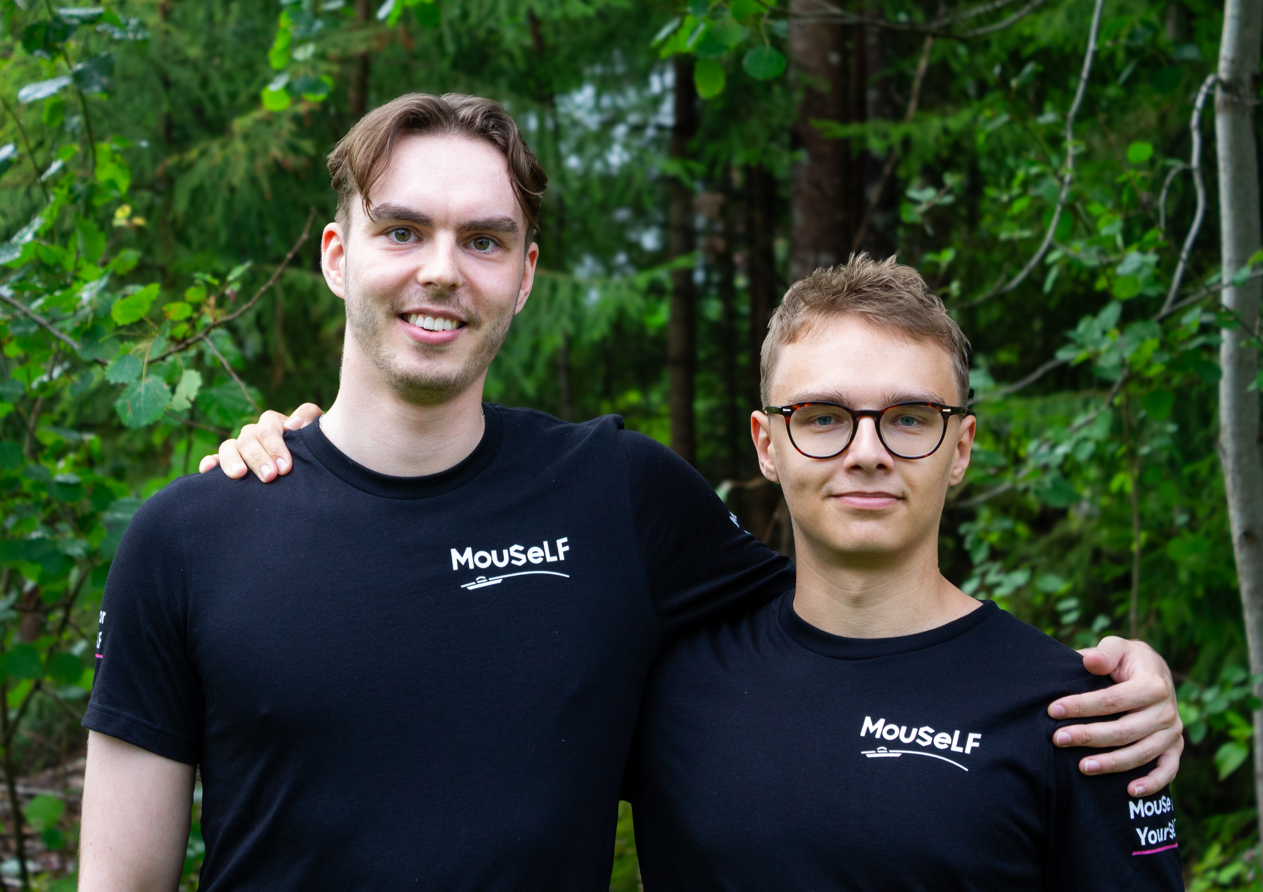 Two young men standing outdoors in a forested area, wearing black t-shirts with the word 'Mouself' printed on them. One has his arm around the other's shoulder and both are smiling.
