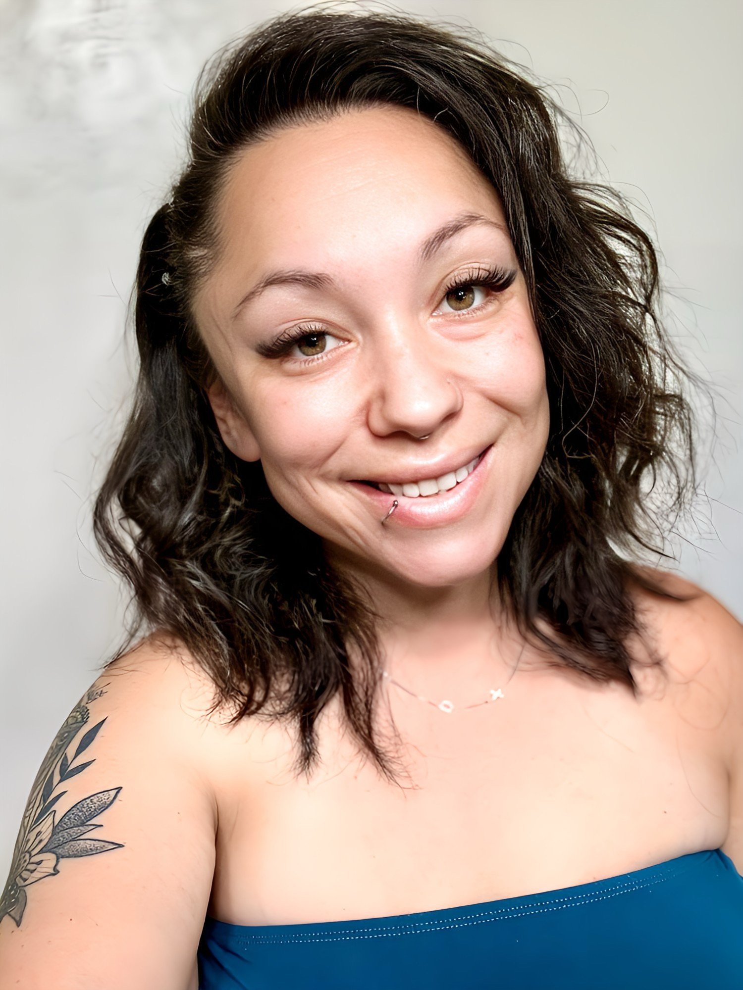 A young woman with curly dark hair, light skin, and tattoos on her shoulder and arm, smiling and looking at the camera, wearing a strapless blue top, in front of a plain background.