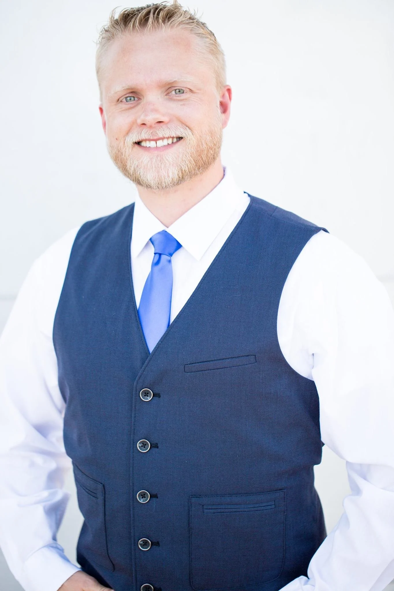 A man with light hair and beard smiling at the camera, wearing a white shirt, blue vest, and blue tie against a plain white background.
