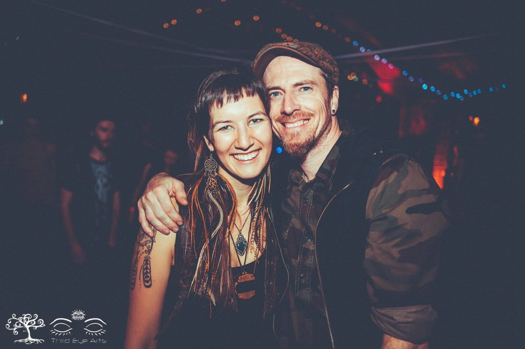 A happy woman and man, both smiling, are standing close together with the woman's arm around the man's shoulder. They appear to be in a dimly lit venue with colorful string lights hanging from the ceiling.