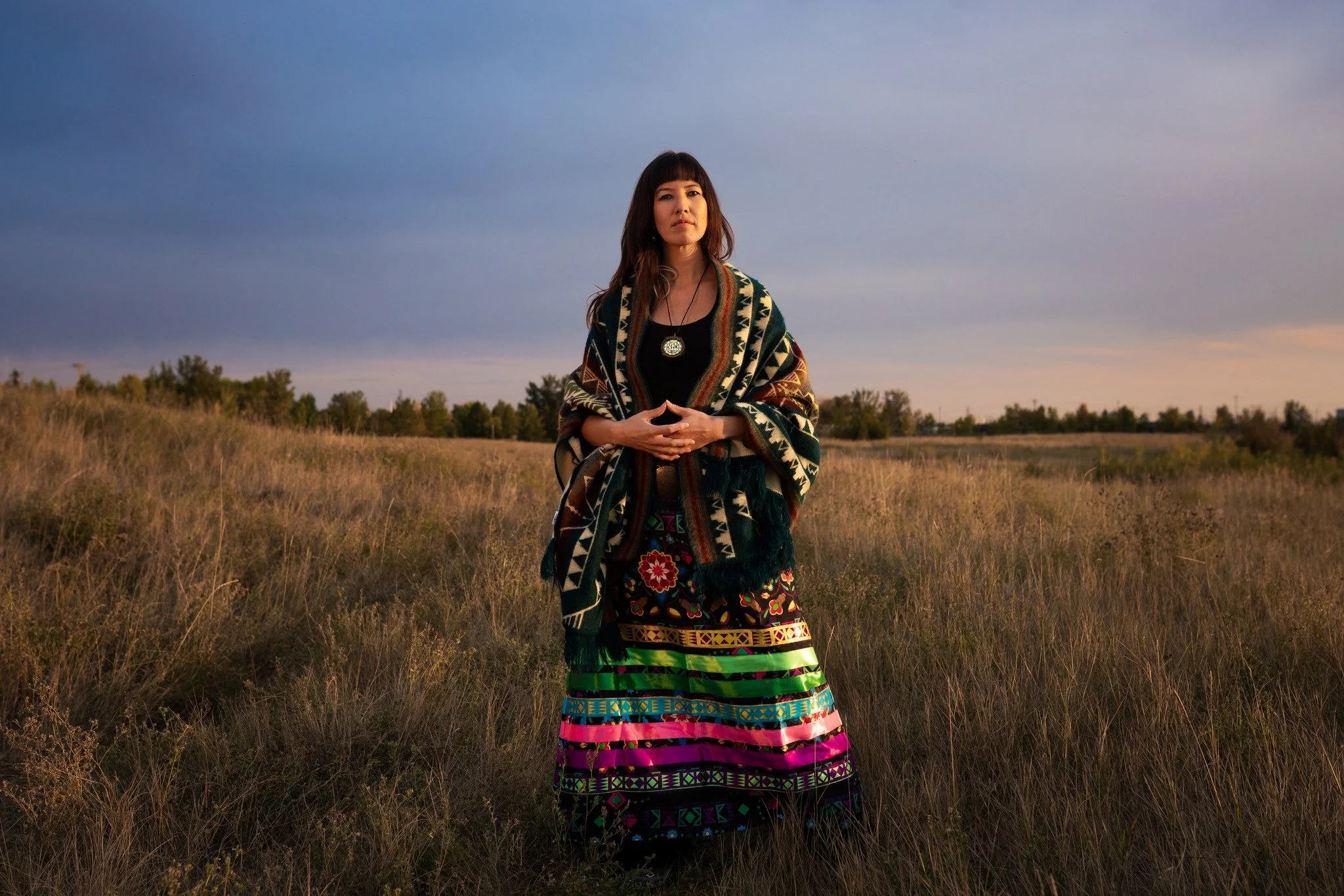 Kylee Dawn standing outdoors in a field with tall grass, wearing a colorful, patterned shawl and a long skirt with vibrant patterns, under a sky with clouds at sunset.
