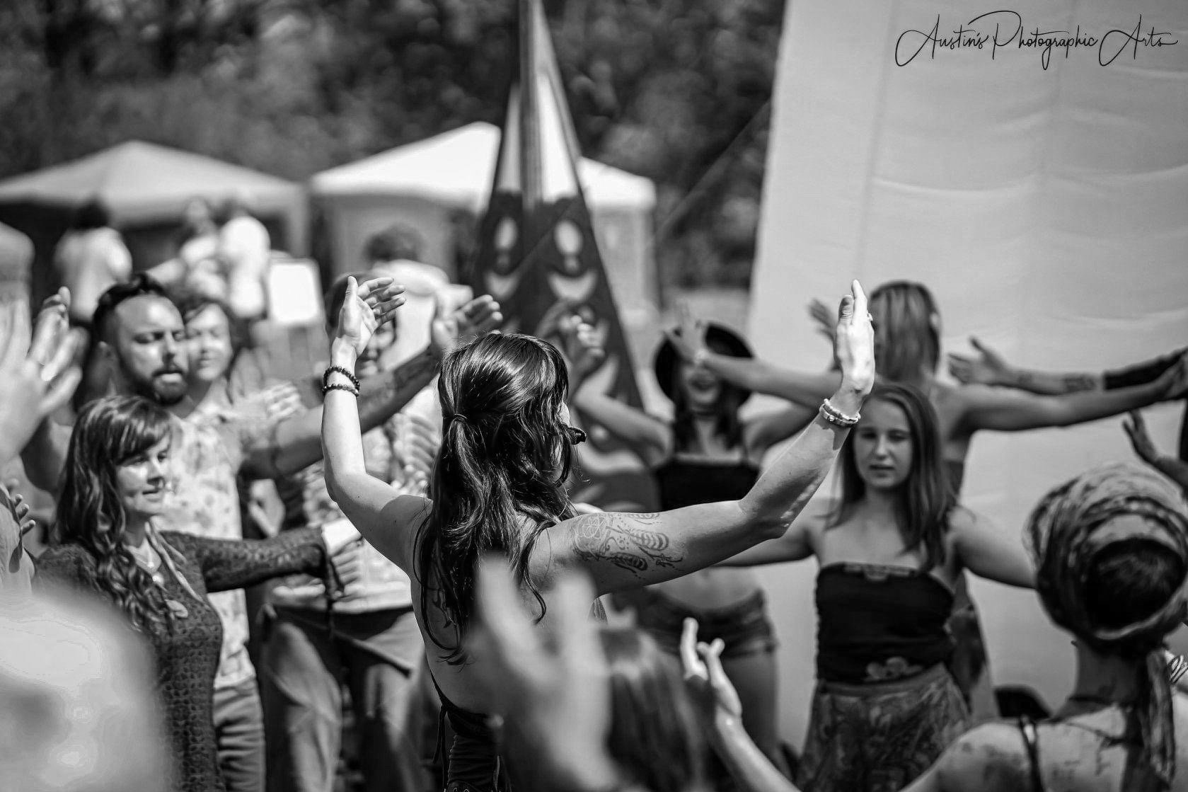 A group of people participating in an outdoor event, with some raising their arms in a dance or celebration. A woman with long hair and tattoos is at the center, with others extending their arms around her. Tents and flags are visible in the backgrou