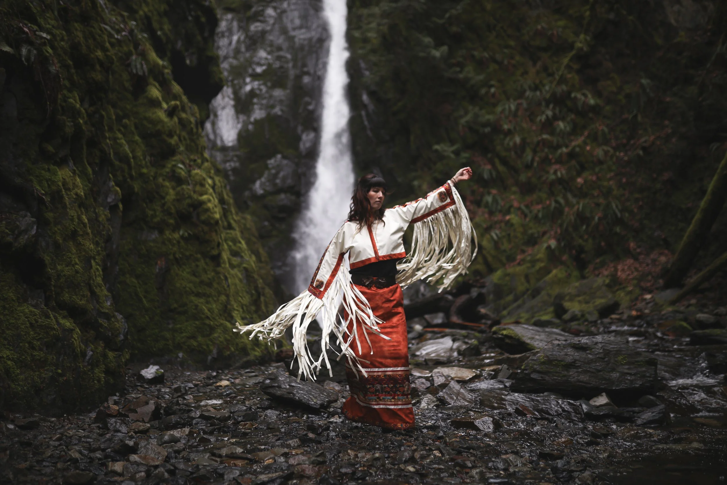 Kylee Dawn dressed in traditional Native American clothing stands in a stream near a waterfall surrounded by moss-covered rocks and dense greenery.