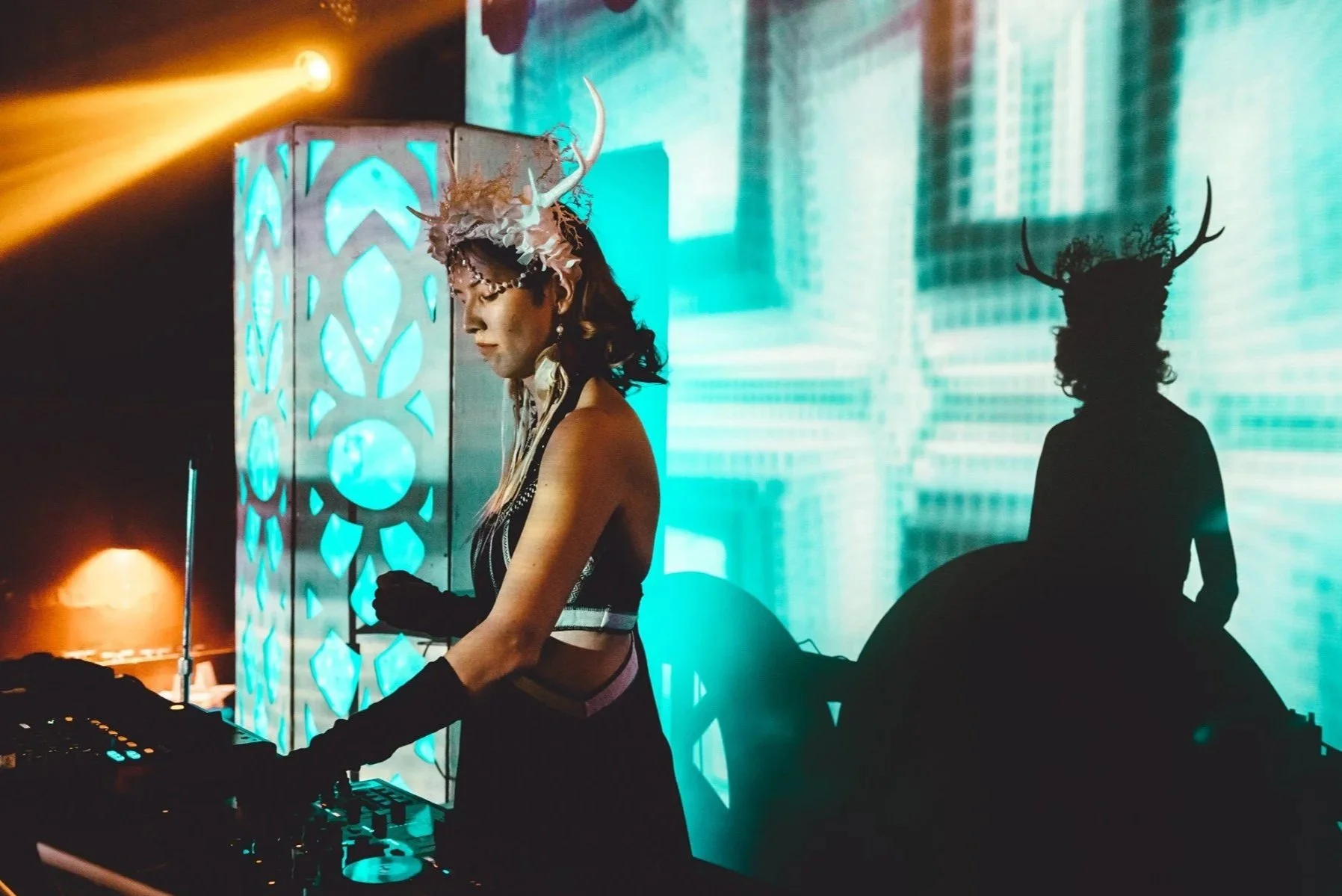 DJ Kylee Dawn wearing a headpiece with antlers and feathers, standing in front of a decorative panel with blue lights, at a music event. Her shadow, with antlers, is cast on a large LED screen behind her.