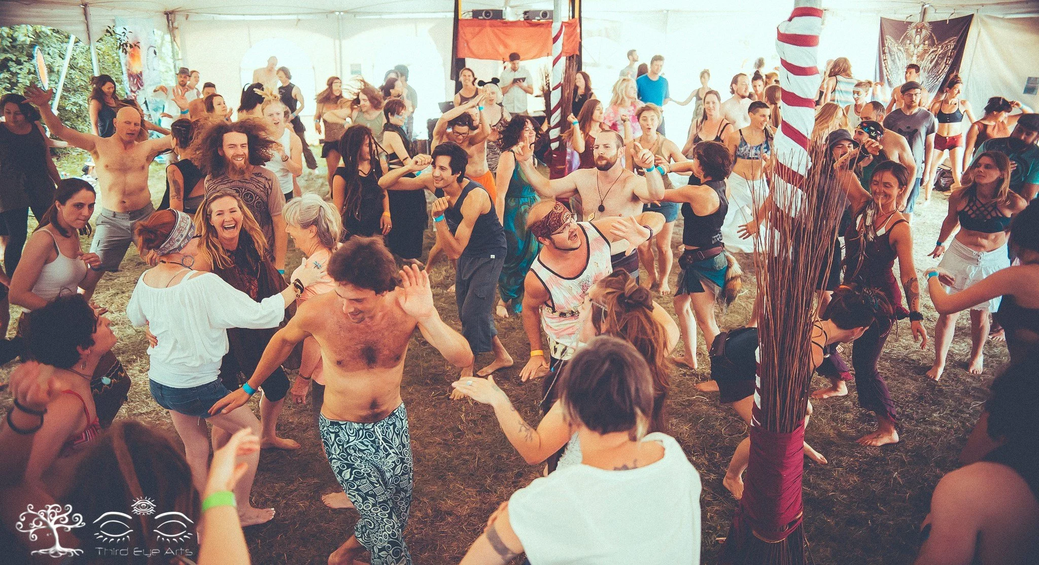 People dancing at Souluna/Inshala and having fun inside a large tent at a festival, with some wearing casual clothing and others in costumes, and a large candy cane prop in the background.
