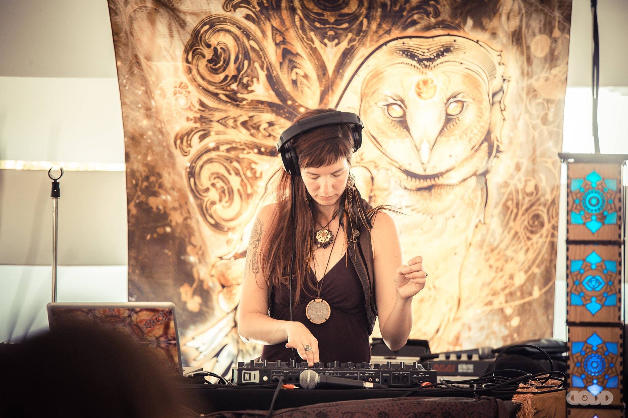 A woman with long brown hair and tattoos on her arm wears headphones and DJ equipment while performing in front of a detailed owl-themed tapestry with swirling designs.