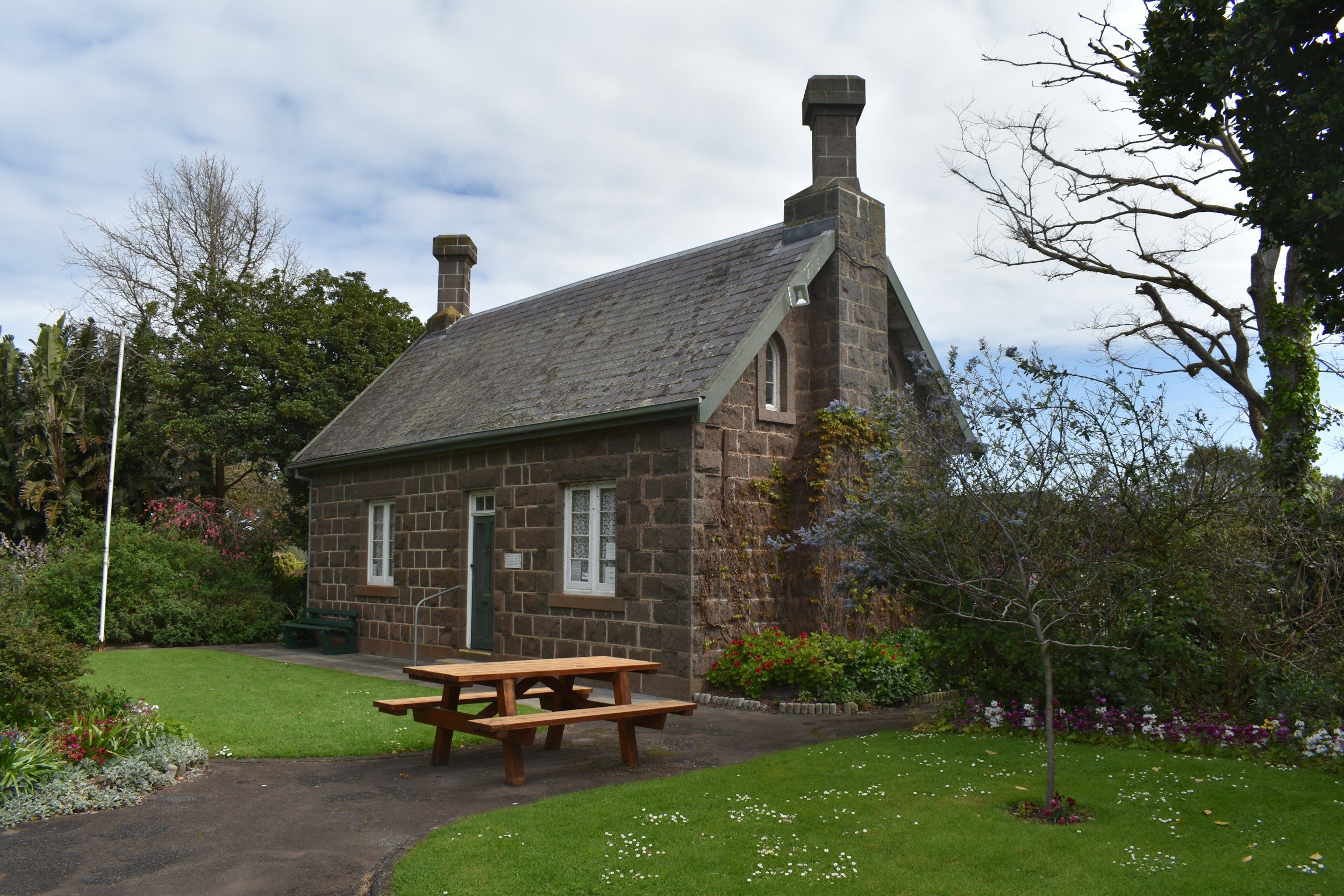 A stone house with a steep roof and two chimneys, surrounded by a garden with trees and flowers, a picnic table in the front, and a pathway leading to the house.
