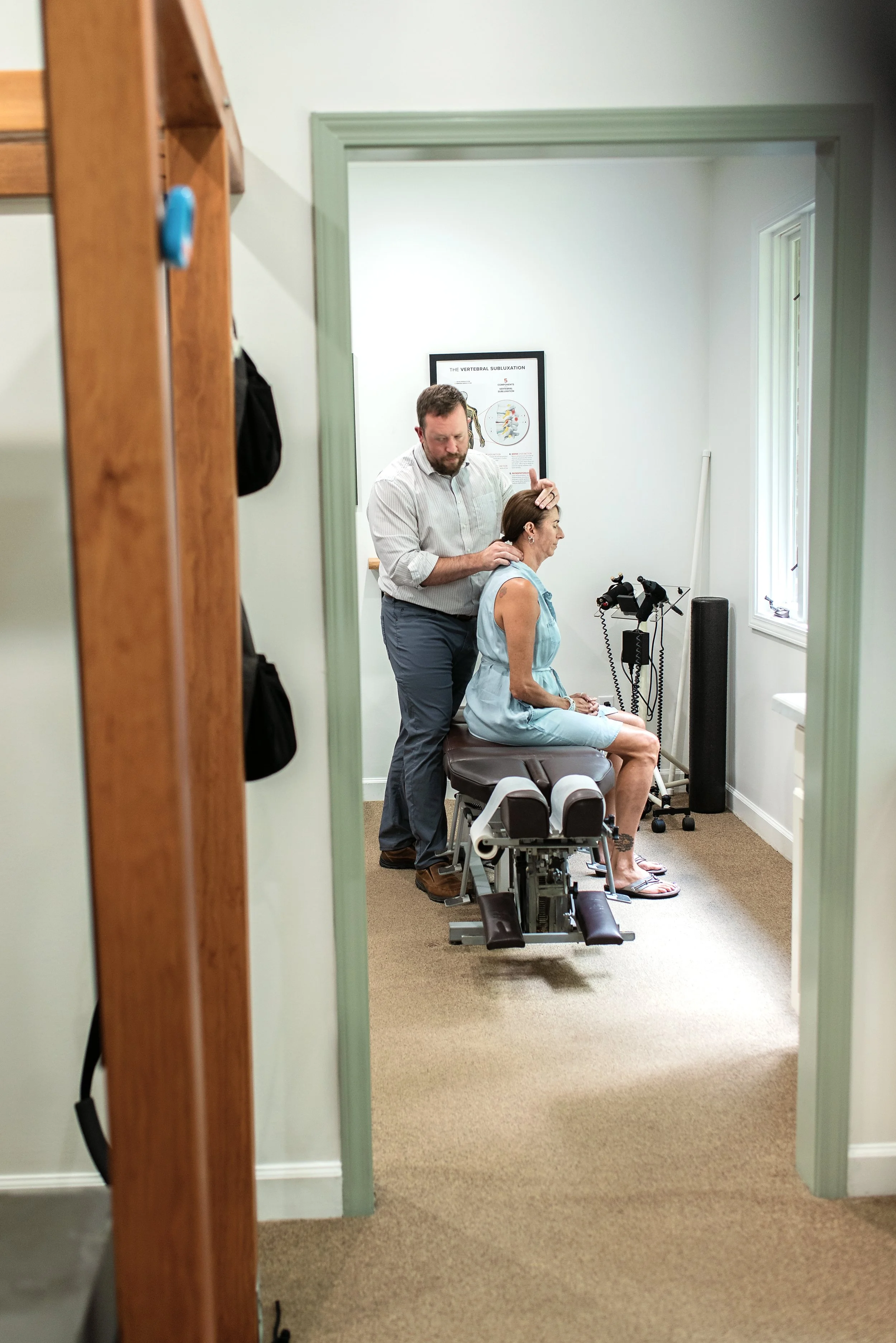 Dr Combs is examining an elderly woman seated on an adjusting table in a Combs Chiropractic & Wellness, with Chiropractic Biophysics traction equipment nearby and a poster about the vertebral subluxation on the wall.